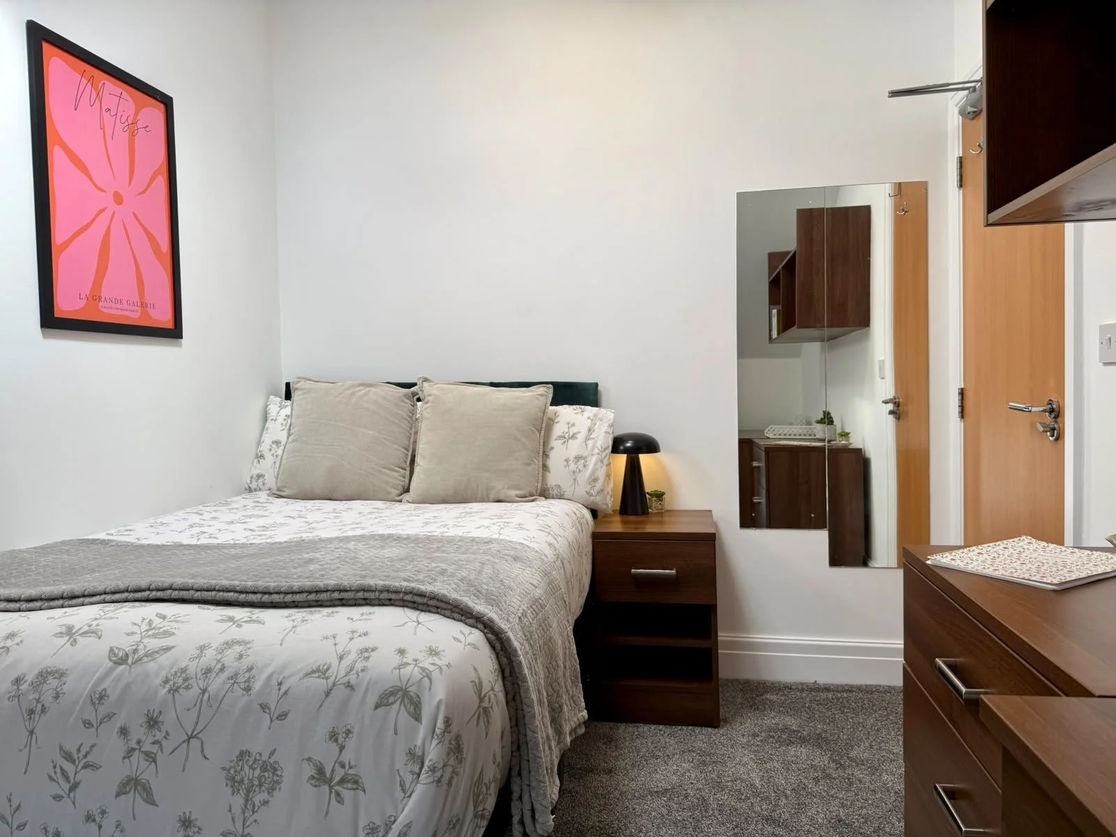 A bedroom with a bed covered in a floral-patterned white and beige quilt, topped with beige pillows. A wooden nightstand with a black lamp and small plant is beside the bed. A mirror and a wooden shelf are on the wall, and a framed pink artwork hangs