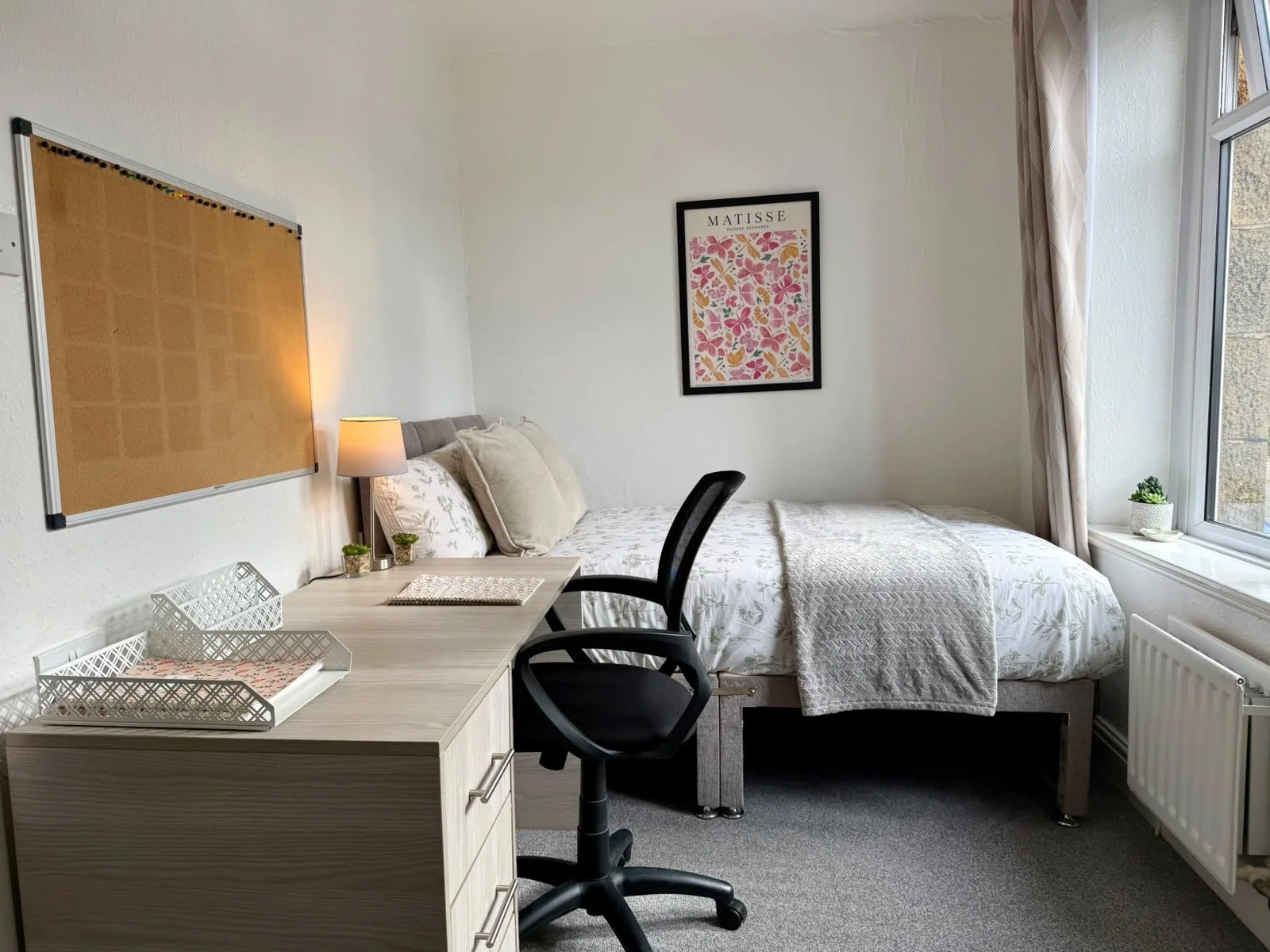 A bedroom with a bed near a window, a desk with a chair, and wall decor.