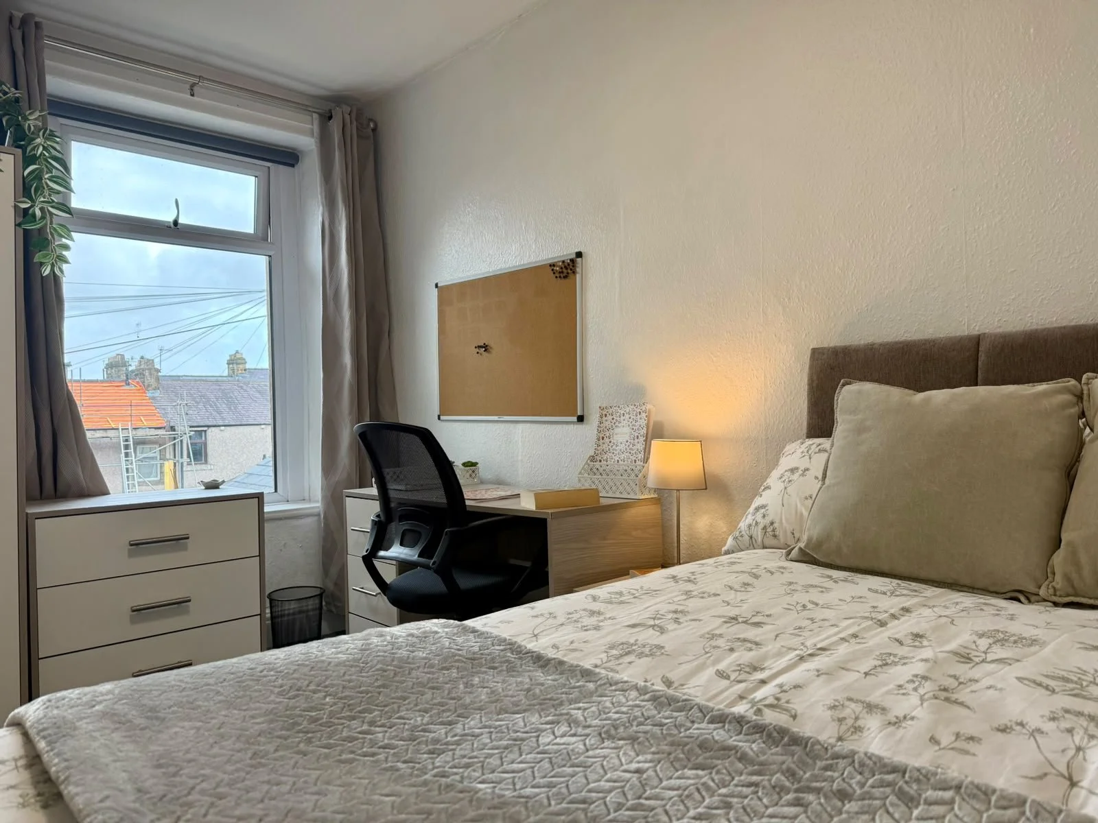 A bedroom with a bed, pillows, a desk with a chair, a small lamp, a window with curtains, and a dresser, with a view of rooftops and chimneys outside.