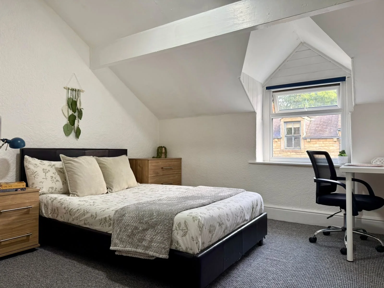 A bedroom with a bed, pillows, a wooden dresser, a black office chair, a white desk, and a window showing a brick building outside.