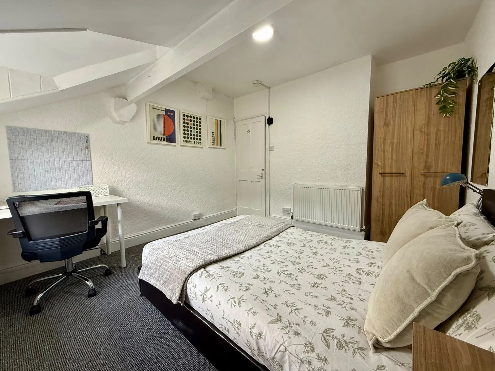 A bedroom with a bed, pillows, a wooden wardrobe, a white desk with a black ergonomic chair, wall decorations, and a radiator.