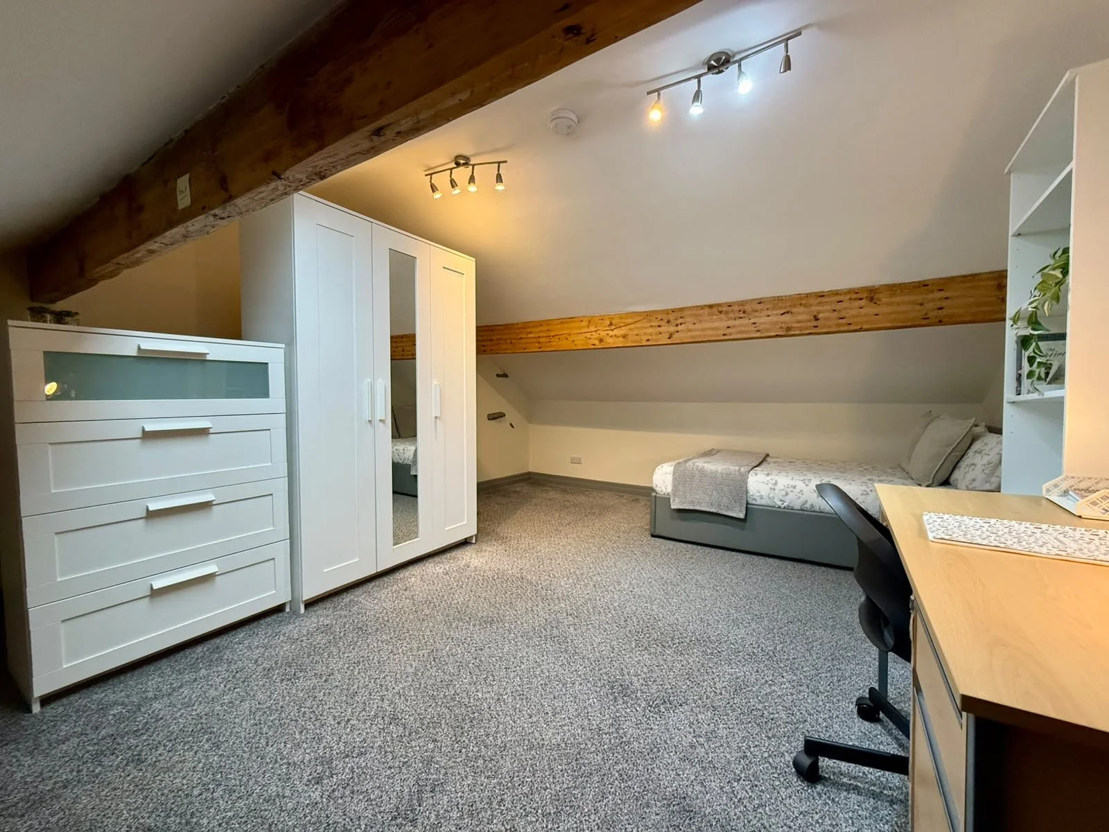 A cozy attic room with a single bed, white dresser, wardrobe with mirror, desk and chair, carpeted floor, and wooden beams on the ceiling, illuminated by ceiling tracks of lights.