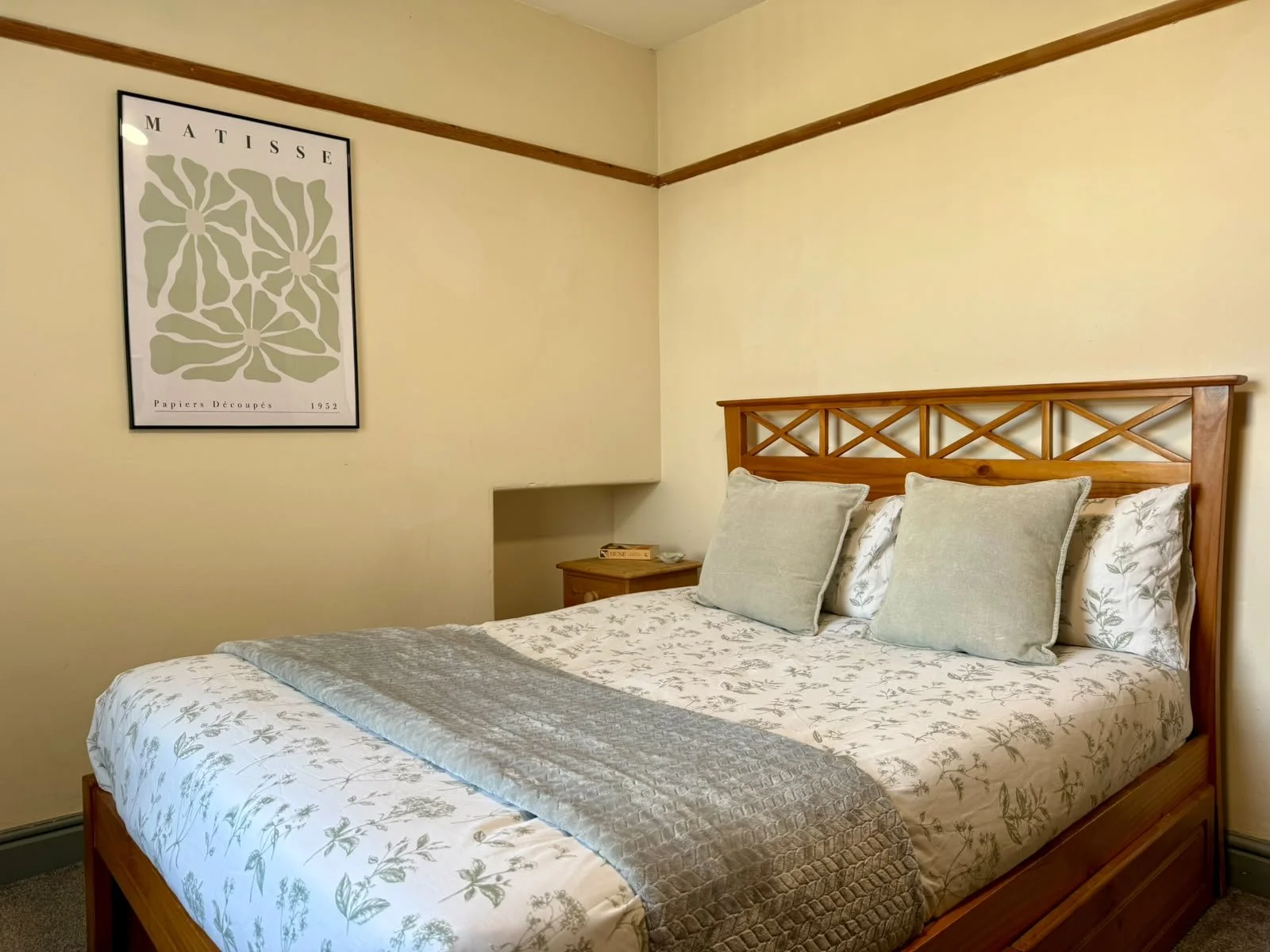 A cozy bedroom with a wooden bed frame, white floral bedding, and gray pillows. A nightstand sits beside the bed with a small box on top. A framed flower print hangs on the wall above.