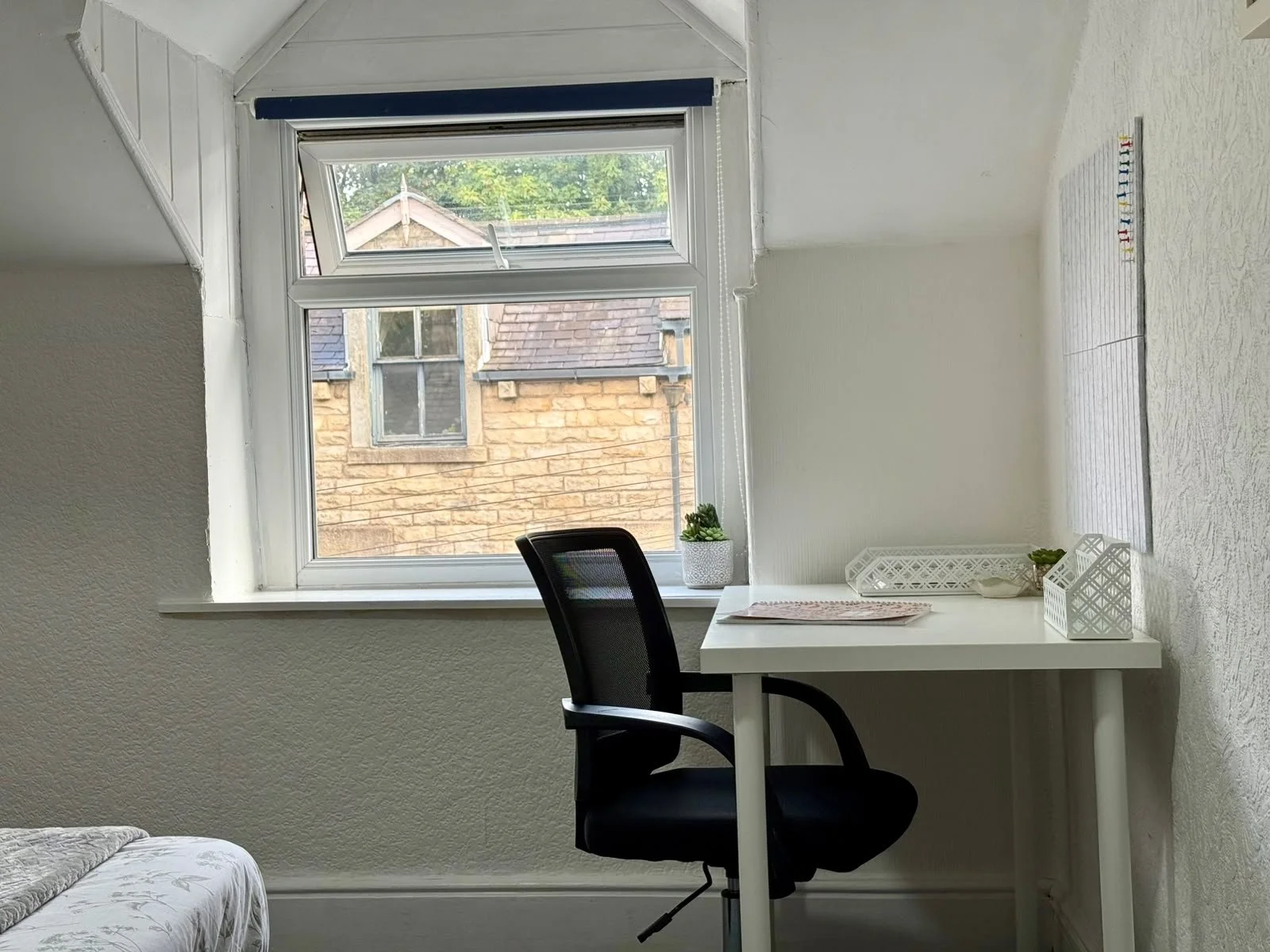 A small white desk with a black office chair in front of a window. The desk has a potted plant, a tray, and a decorative container. The window shows a view of neighboring brick and stone buildings and trees.