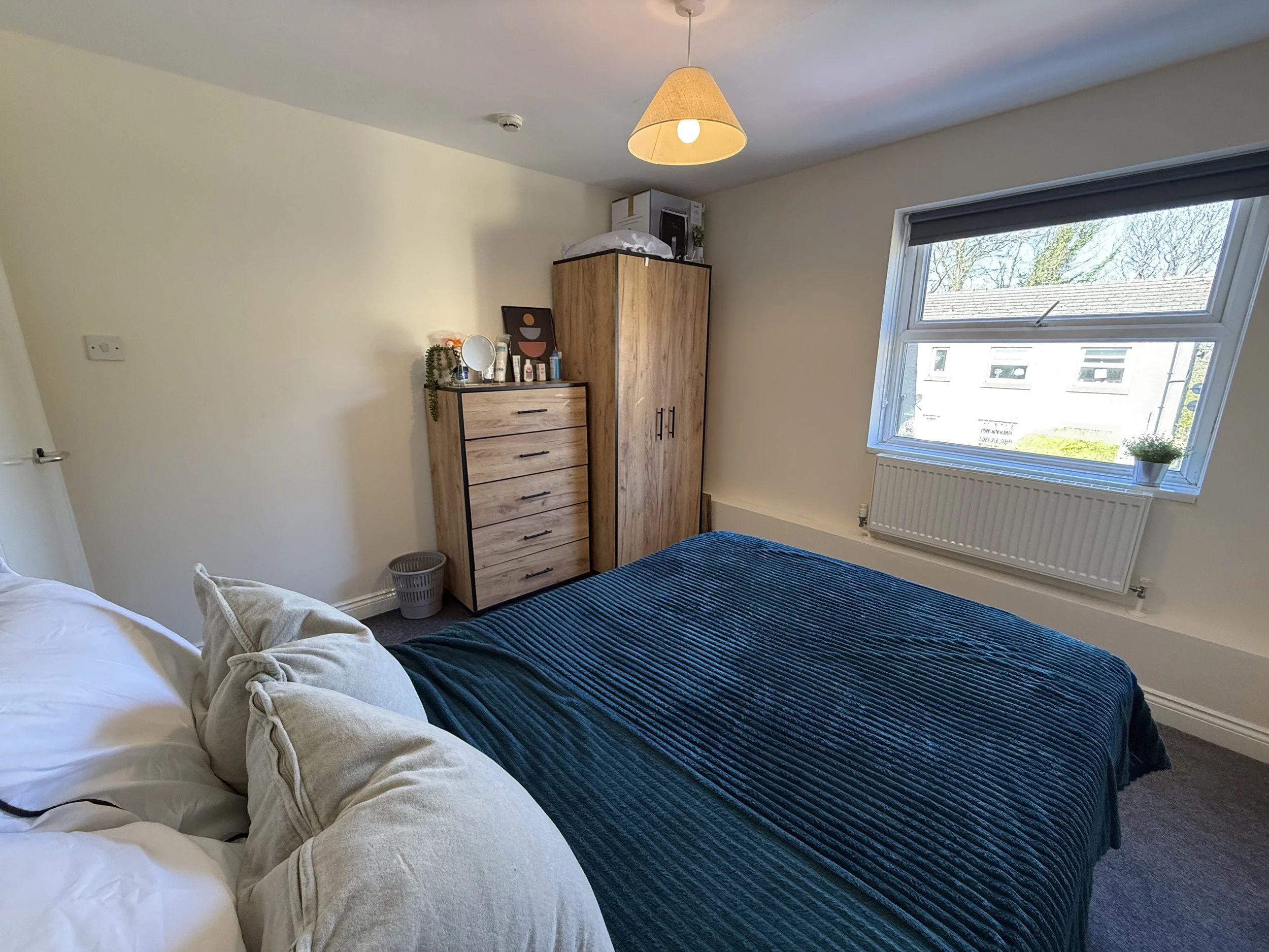 A bedroom with a bed covered in white and dark blue bedding, a wooden wardrobe, a matching dresser with personal items, a window with a view of neighboring buildings, and a ceiling light