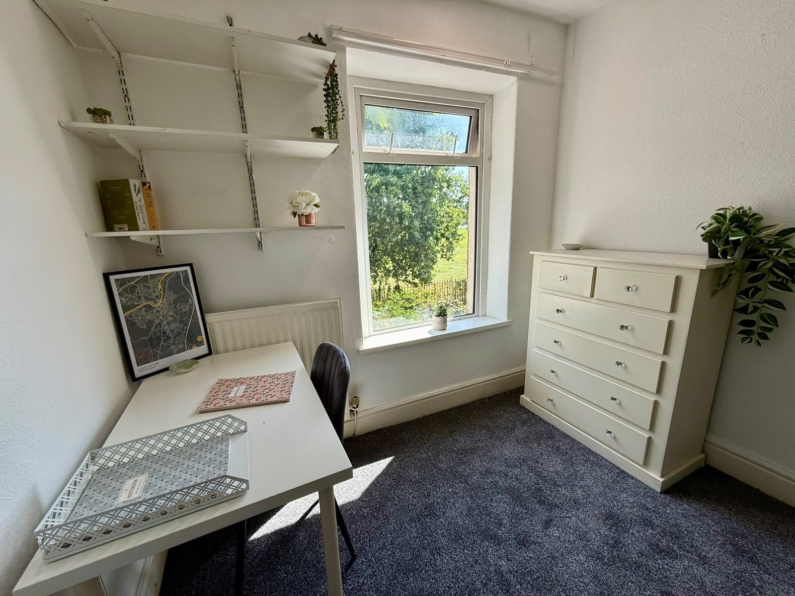 A small home office with a white desk, black chair, open window with a view of green trees, white chest of drawers, and white wall shelves decorated with plants and books.