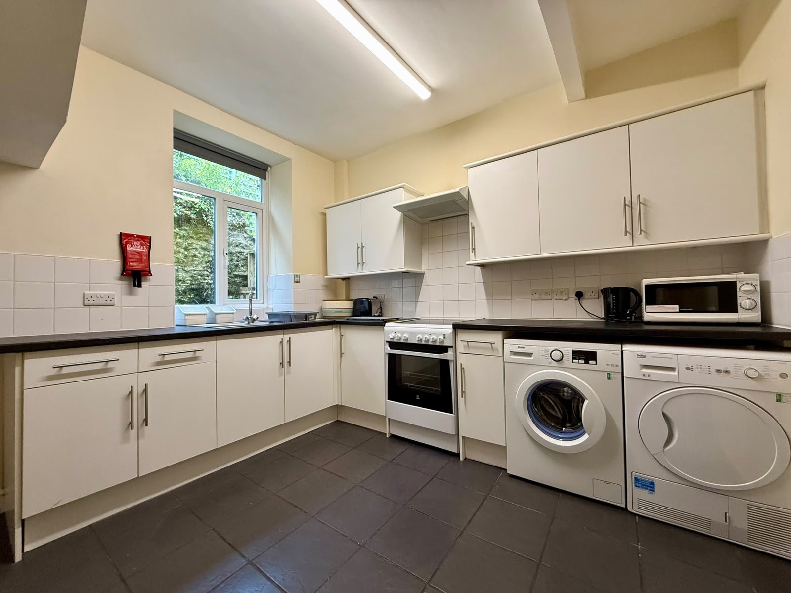 Kitchen with white cabinets, black countertops, washing machine, dryer, oven, microwave, kettle, and sink near a window