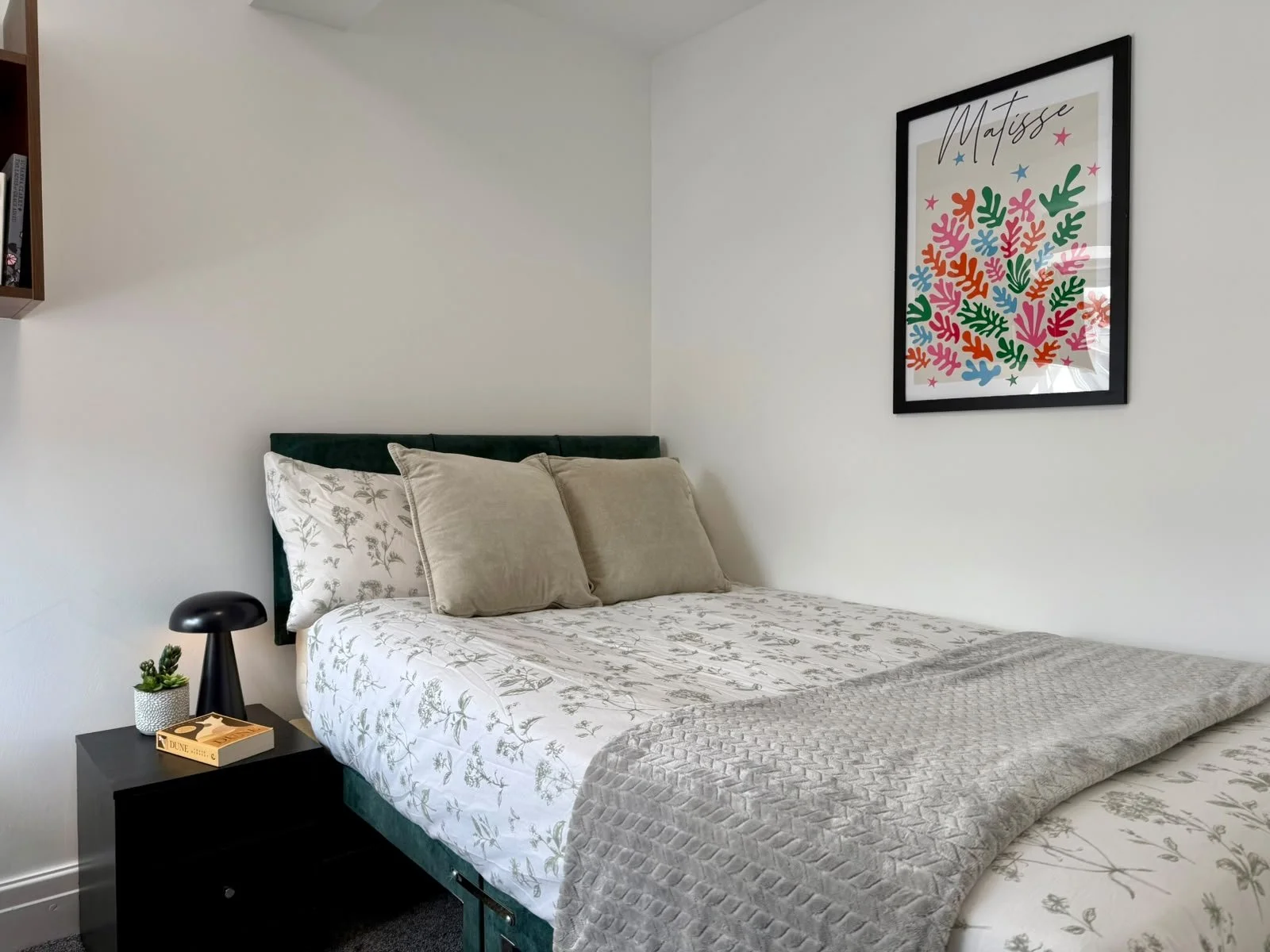 A bedroom with a bed that has floral-patterned sheets and beige pillows. A black nightstand with a small plant, a book, and a black lamp, and a colorful framed art print on the white wall.
