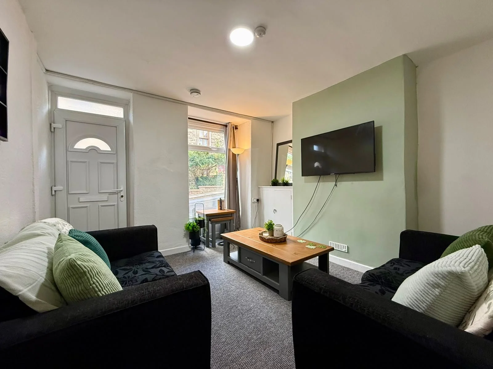 Living room with two black sofas with green and white pillows, wooden coffee table, mounted flat-screen TV on a light green wall, window with curtains, white door with window, potted plants, and decorative items.