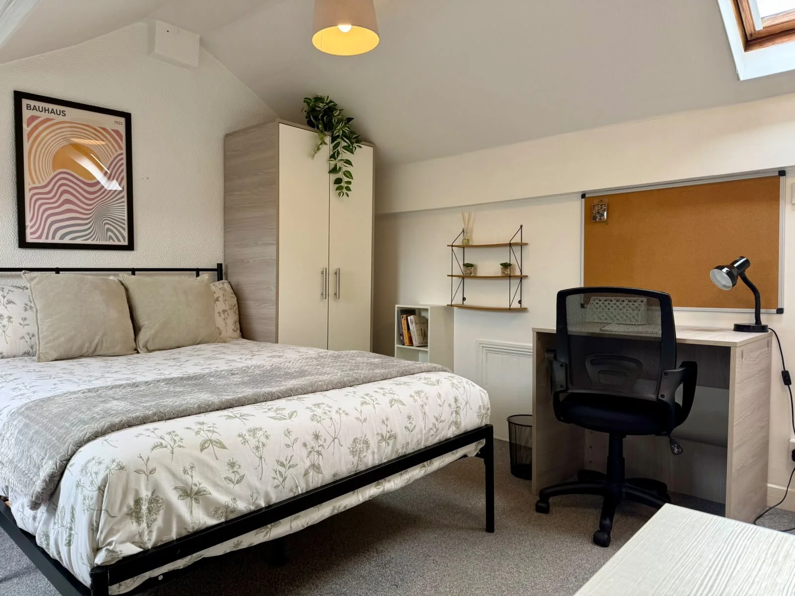 A bedroom with a bed, pillows, a desk with a chair, a lamp, a cork bulletin board, some shelves, and a wardrobe. The room has a sloped ceiling with a skylight and a framed artwork on the wall.