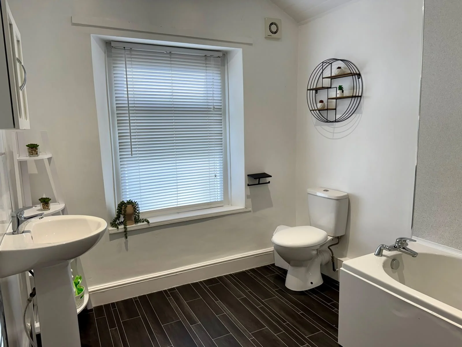 A bathroom with a white sink, a toilet, a bathtub, and decorative shelves on white walls, with a window with blinds and potted plants.