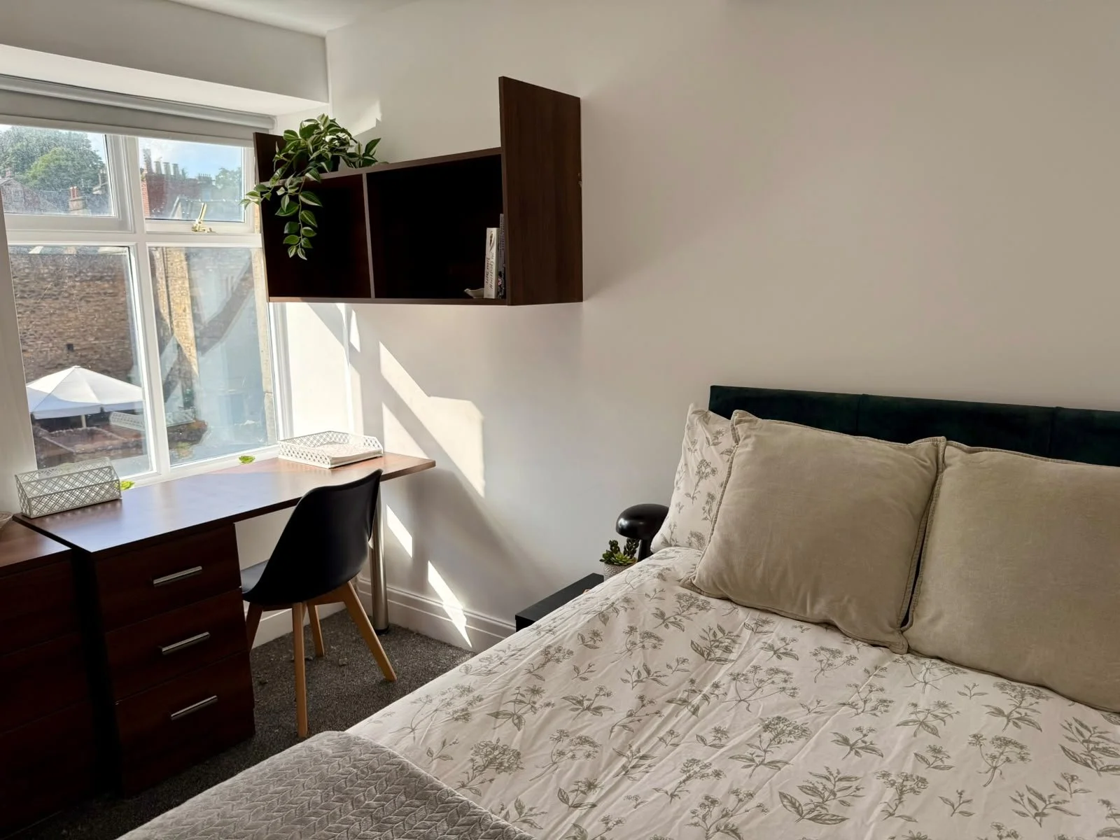 A bedroom with a window overlooking rooftops, a desk with a chair, a wall shelf with a plant and books, and a bed with floral bedding and beige pillows.