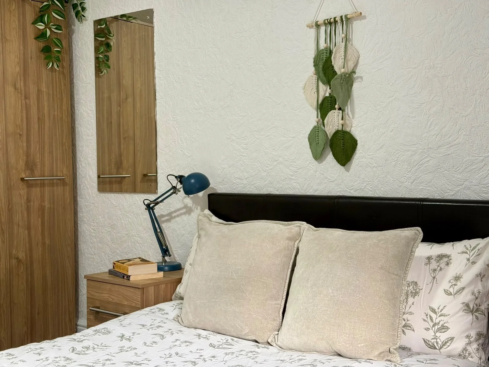 Bedroom with a bed, beige pillows, a black headboard, a wooden nightstand with a blue desk lamp and books, a closet with wooden doors, a mirror, a wall hanging decorated with green and white leaves, and a white textured wall.