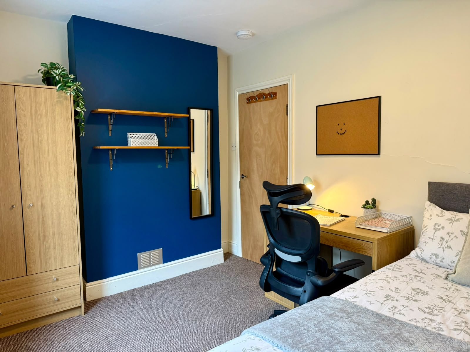 A cozy bedroom corner with a wooden desk, black ergonomic chair, bed with floral bedding, a small potted cactus plant, a bulletin board with a smiley face, and a standing mirror against a white wall. A navy blue accent wall with floating shelves and 