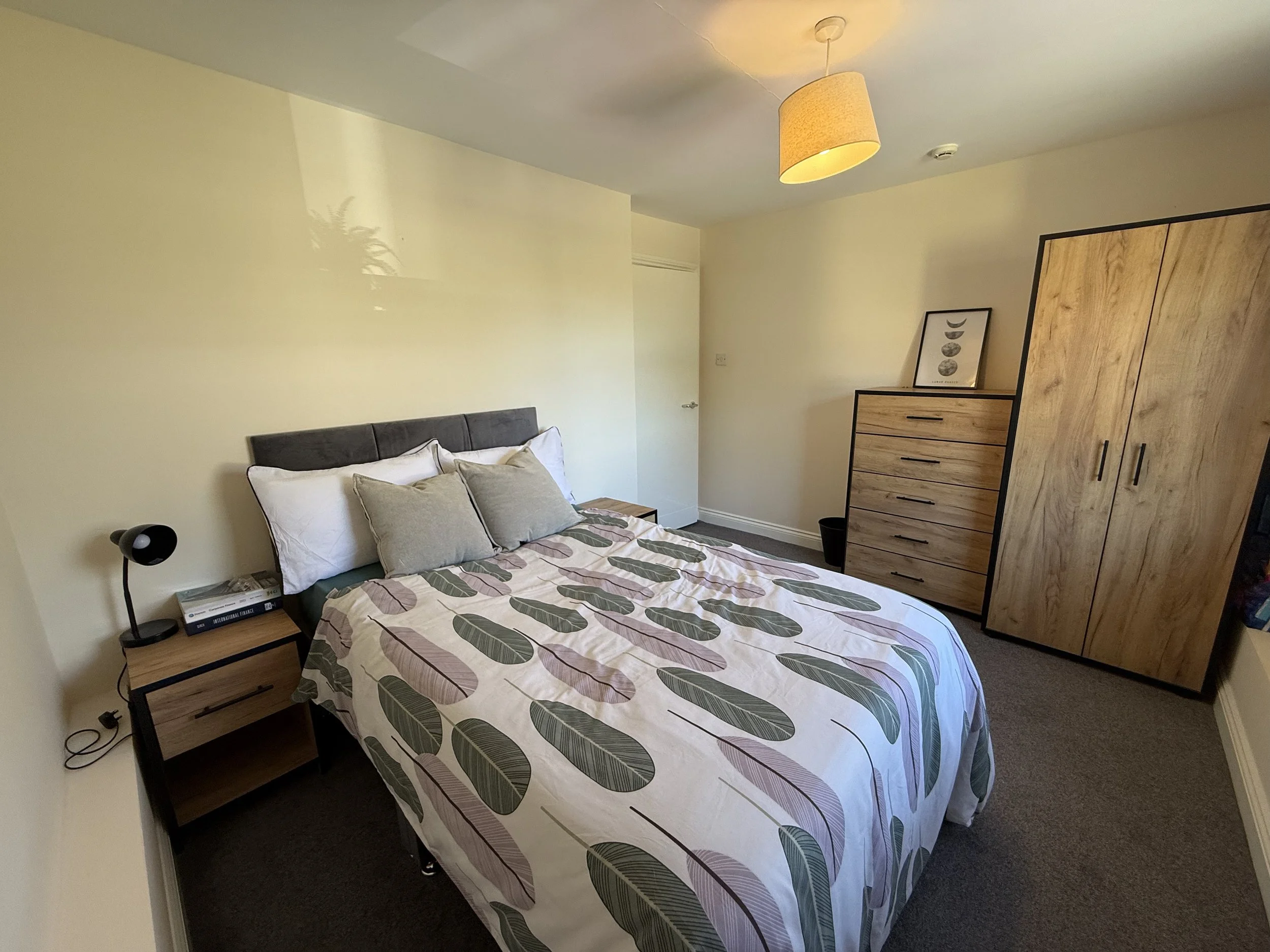 A bedroom with a queen-sized bed, a gray headboard, and a bedsheet with leaf patterns in shades of green, beige, and purple. The bed has four pillows, two white and two gray. To the left of the bed, there is a small wooden nightstand with a black des