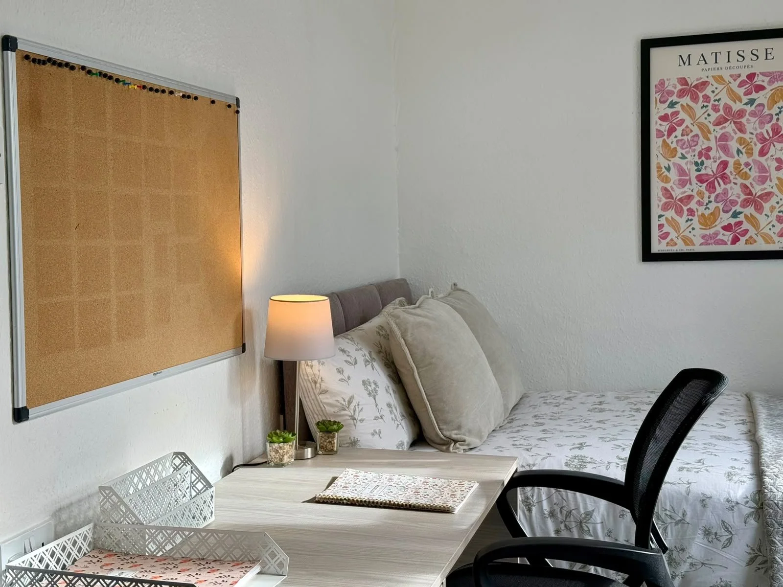 A cozy bedroom with a bed, pillows, a bedside lamp, and wall art, adjacent to a work or study desk with office supplies and a black chair.