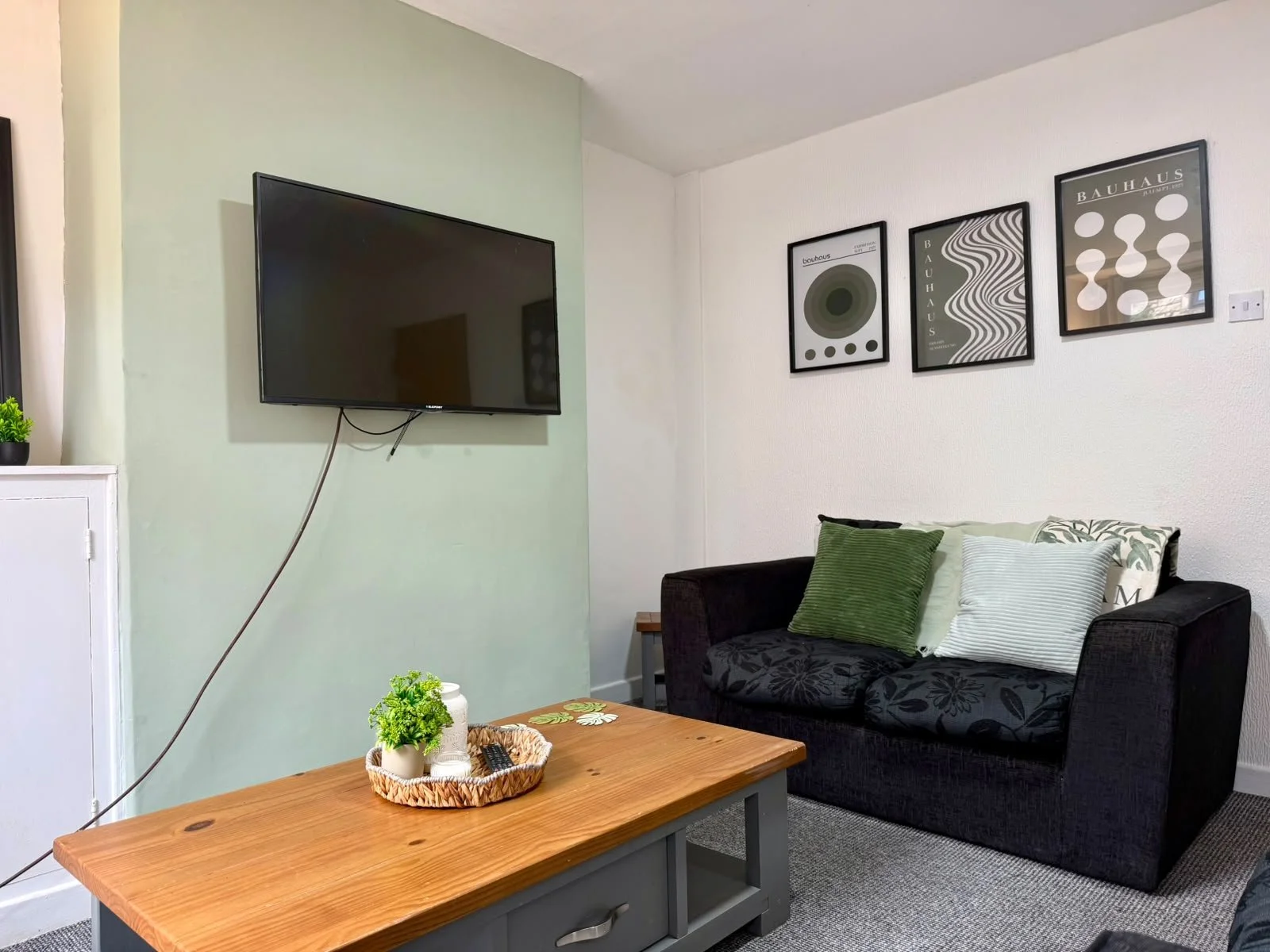 Living room with a wall-mounted TV on a light green wall, a black sofa with green and white cushions, a wooden coffee table with a small potted plant, a remote, and coasters, and framed abstract artwork on the white wall.