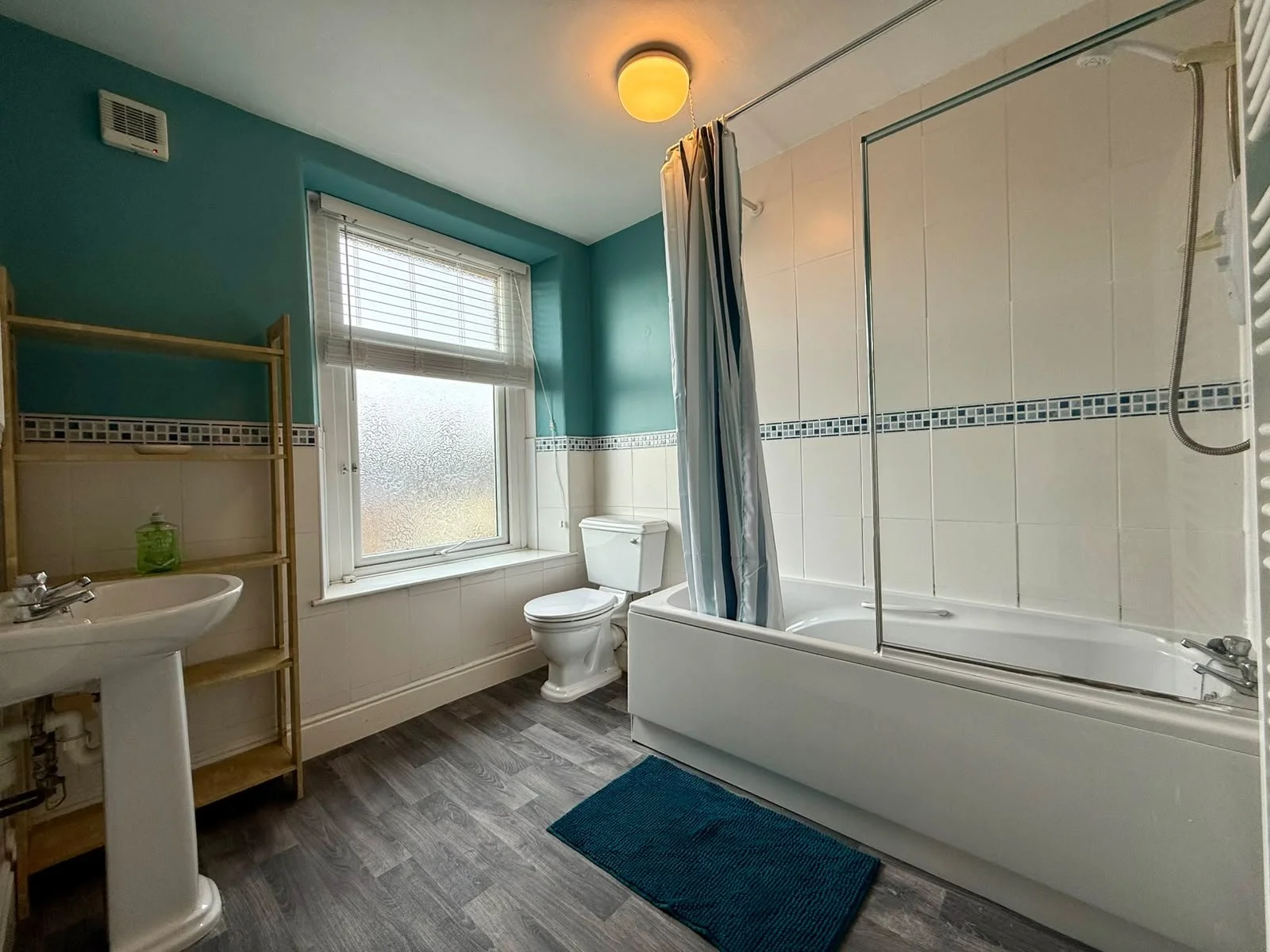 A bathroom with a window, bathtub with shower, toilet, sink, and shelving unit. The walls are painted teal, and the floor has wood-patterned vinyl. The window has blinds, and there is a blue bath mat outside the tub.