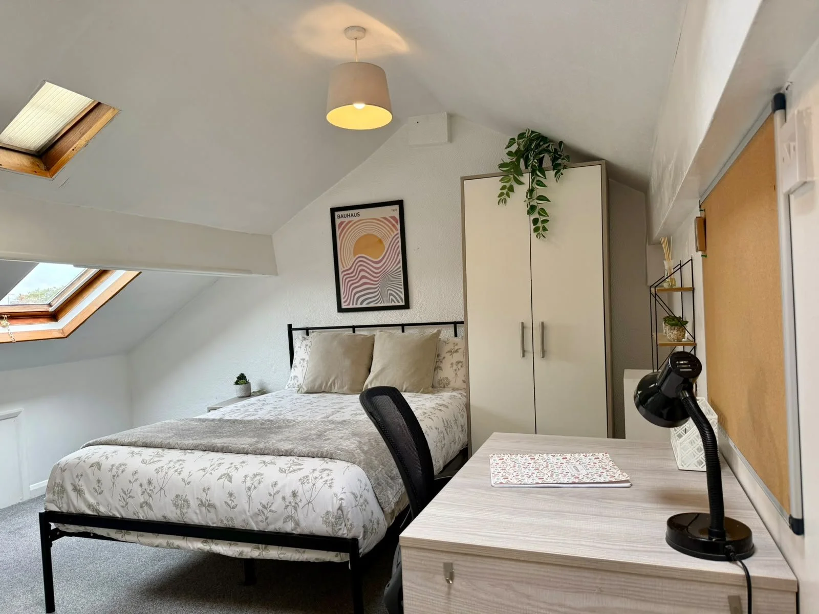 An attic bedroom with a bed, wardrobe, and desk, featuring skylights and a ceiling light. There are various decorative items like a poster, plants, and desk accessories.