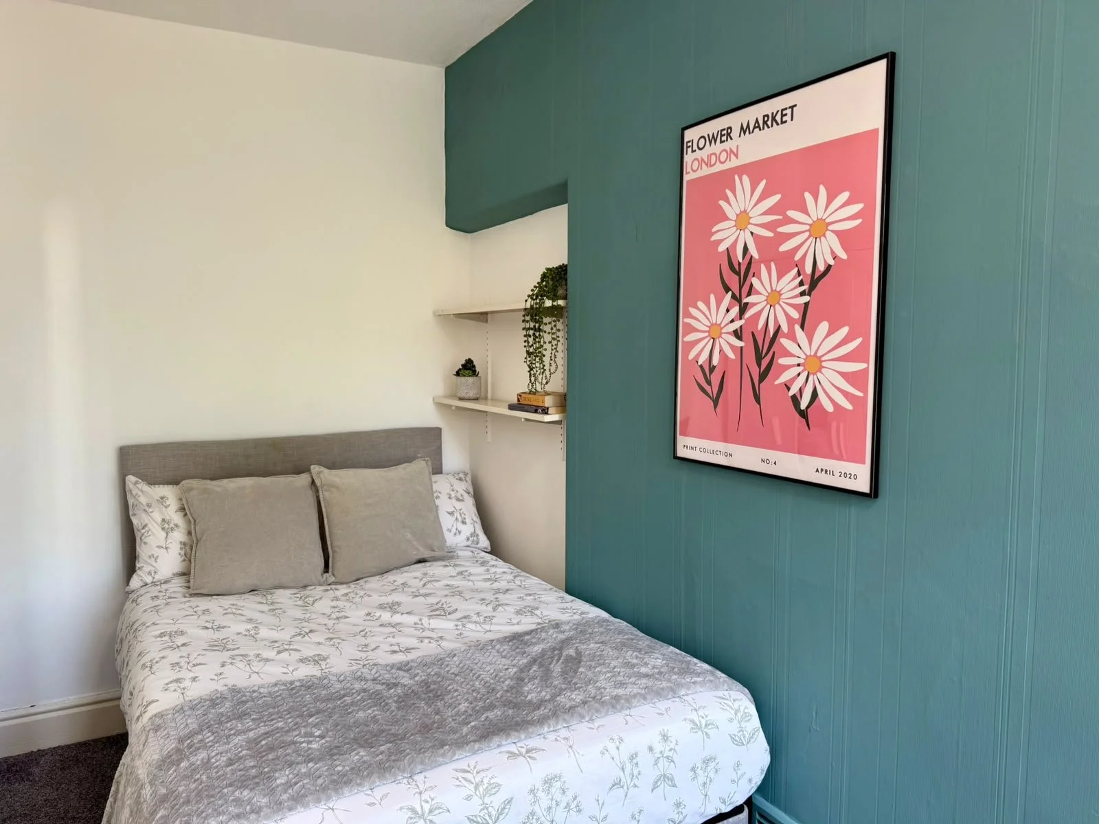 Bedroom with gray bed and beige pillows, floating shelves with plants and books, large framed pink and white daisy flower poster on teal wall.