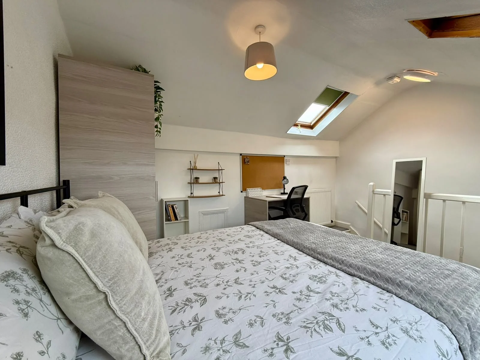 A cozy bedroom with a bed that has floral-patterned bedding and pillows. There is a wooden headboard, a small bookshelf, a desk with a chair and lamp, a window with a skylight, and a staircase leading to the lower level. The ceiling has a hanging lig