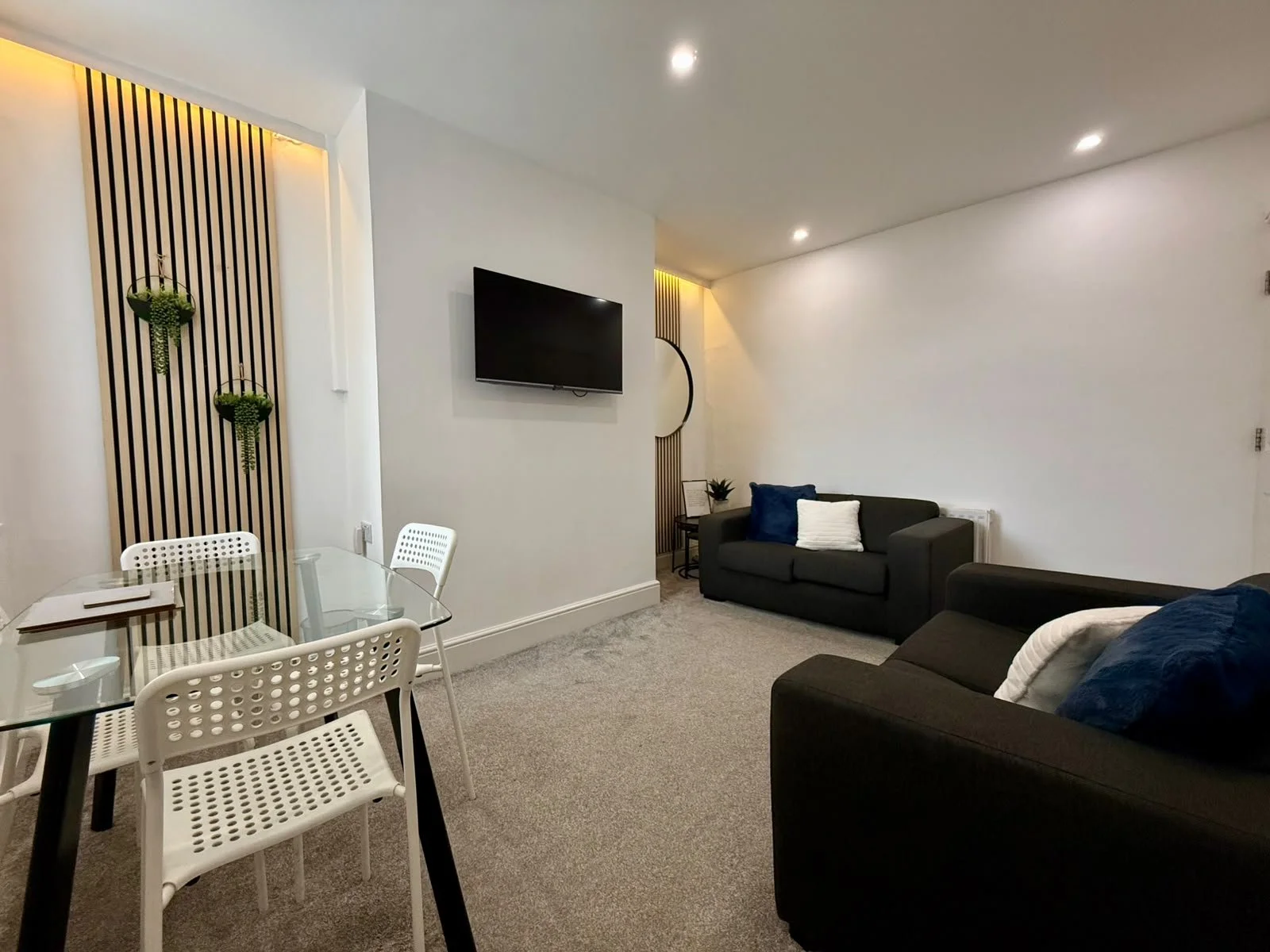Living room with white walls, beige carpet, and modern furniture including two dark sofas with pillows, a glass dining table with white chairs, a wall-mounted TV, and decorative plants, enhanced with warm lighting.