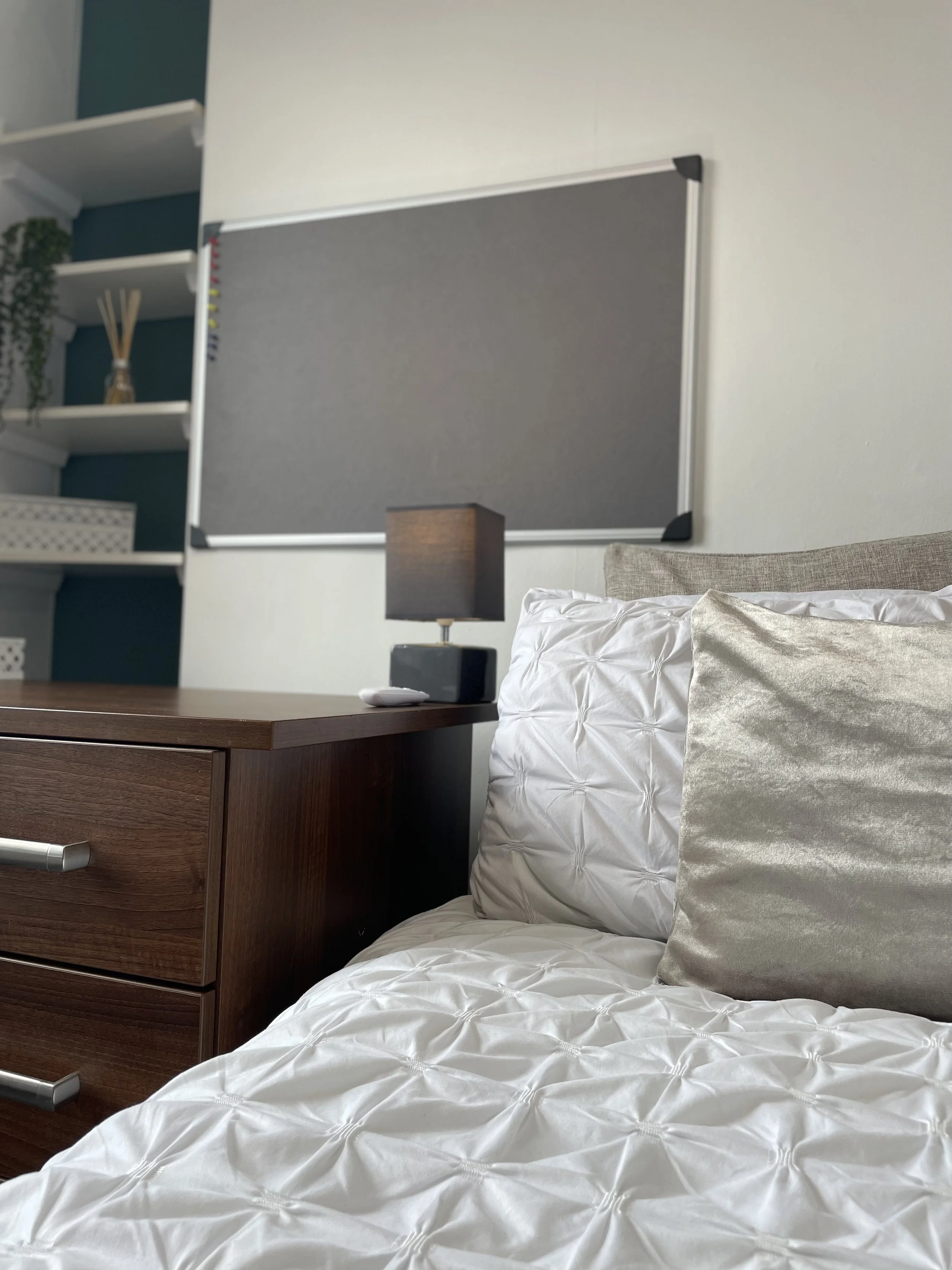 Close-up of a bedroom with a nightstand, table lamp, bed with white bedding and pillows, and a bulletin board on the wall.