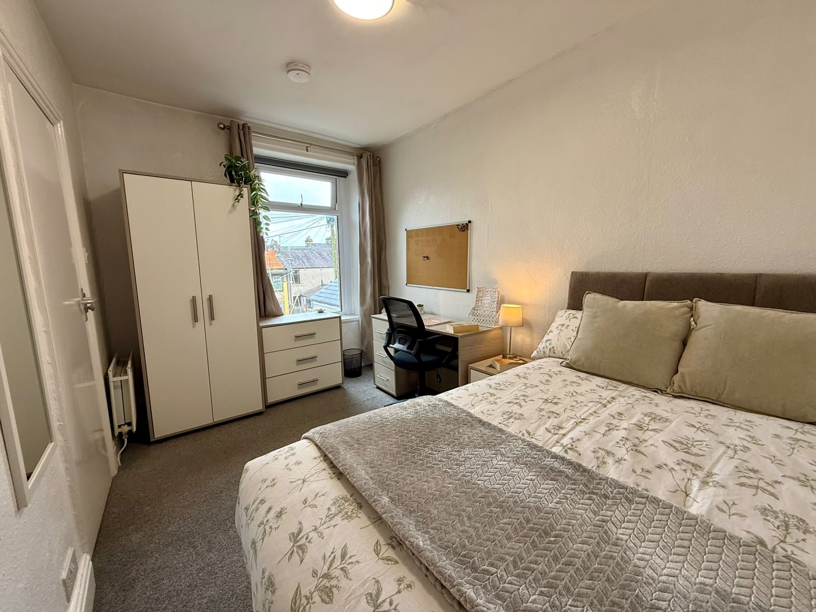 A bedroom with a bed, pillows, a bedside lamp, a desk, a chair, a wardrobe, a window with curtains, a white dresser, and a corkboard on the wall.
