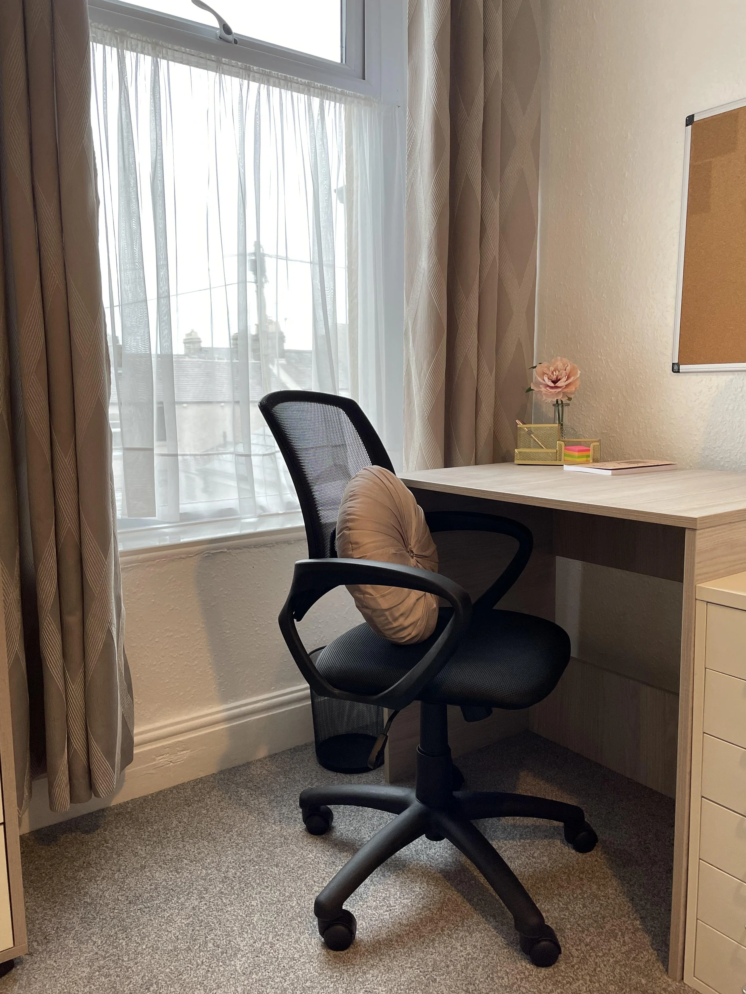 An office space with a desk, a black ergonomic chair with a beige cushion, a window with sheer curtains, and a bulletin board on the wall. The desk has a pink flower in a vase, a pink sticky note pad, and some papers.
