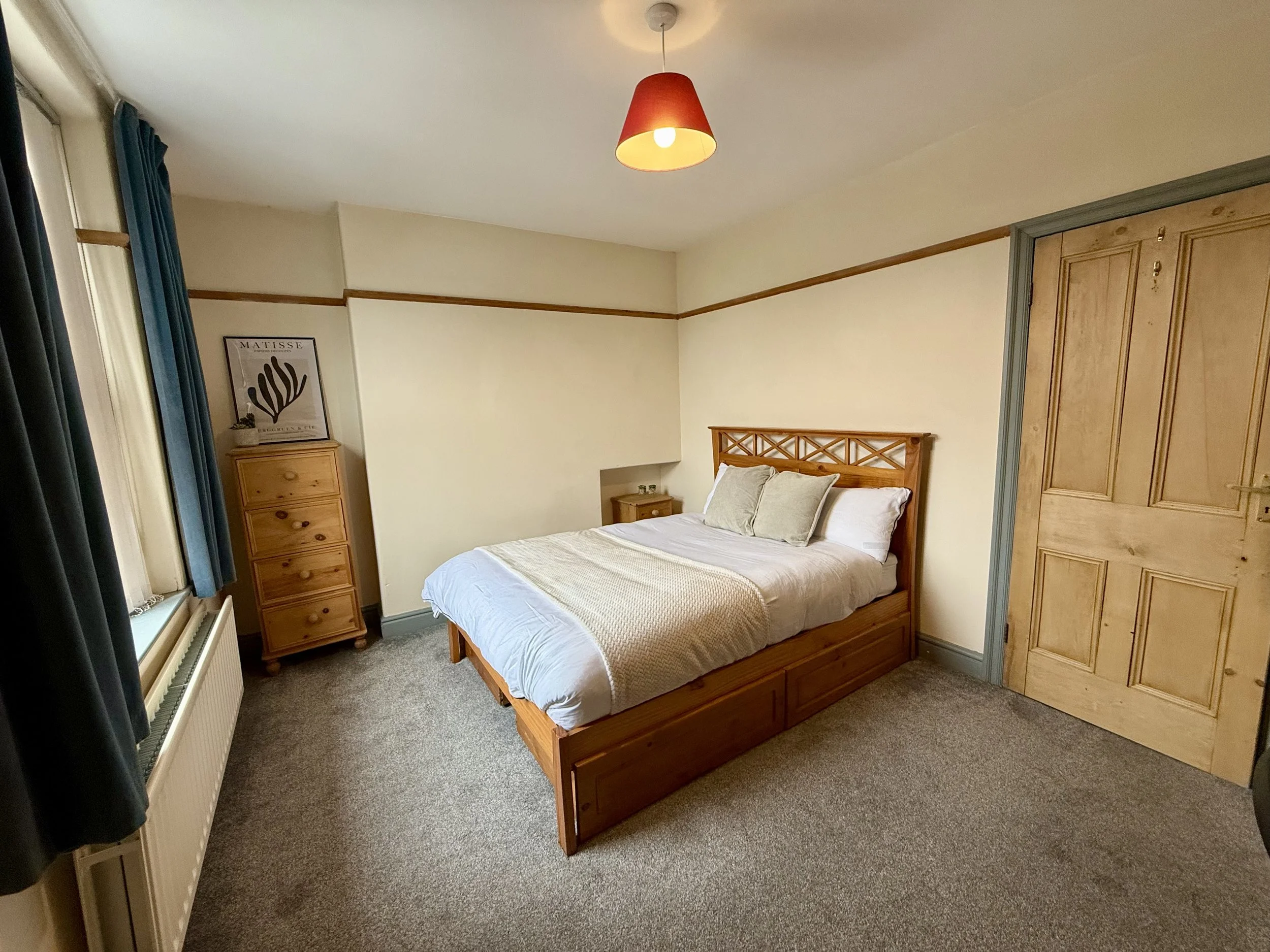 A cozy bedroom with a wooden bed frame, beige bedding, beige pillows, a small wooden bedside table, a wooden dresser, and a window with dark curtains. The walls are painted cream with a wooden trim, and there's a red lampshade hanging from the ceilin