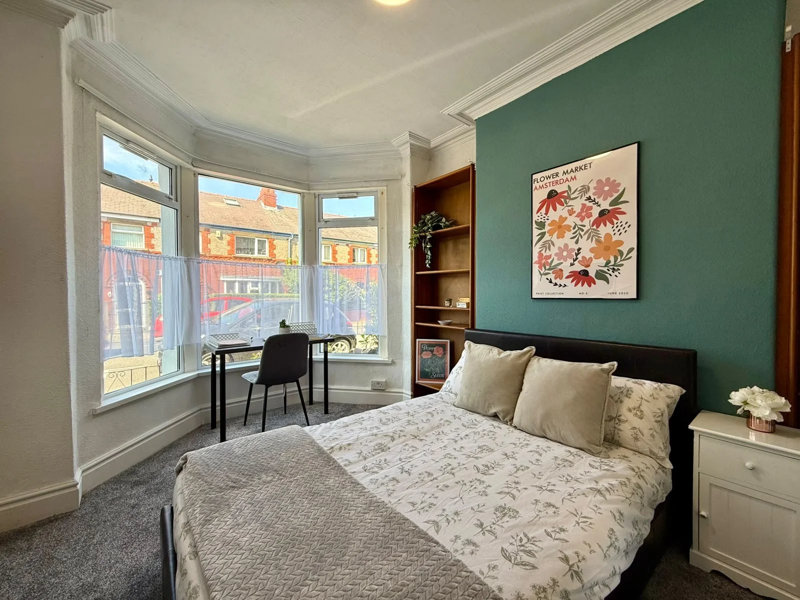 A cozy bedroom with a large bay window, single bed with patterned bedspread and pillows, a nightstand with flowers, a small desk and chair by the window, and a blue accent wall with a floral art print.