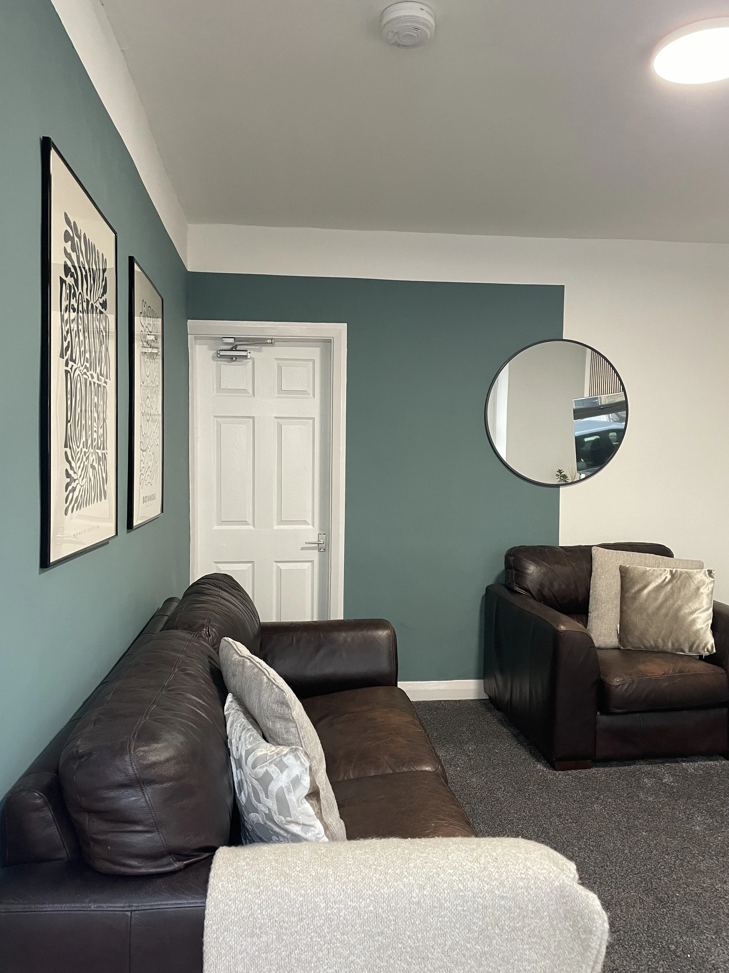 A cozy living room with two dark brown leather sofas, gray throw pillows, wall art, a wall mirror, a door, and ceiling lights.