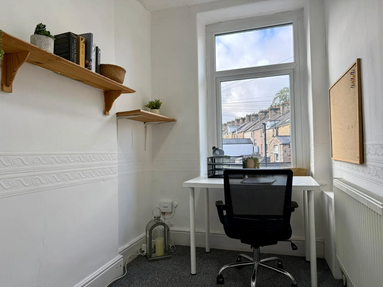 A small home office with a white desk near a large window showing rooftops and sky. A black office chair is positioned at the desk, which has a black pen organizer, a potted plant, and a desk lamp. There are two wooden shelves with books and small de
