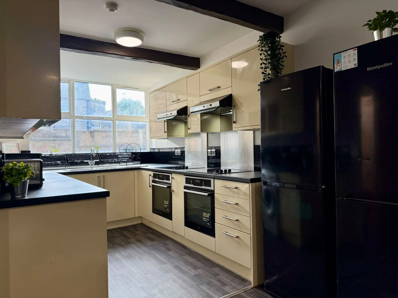 A modern kitchen with cream-colored cabinets, black countertops, and a large window letting in natural light. There are two built-in ovens, a black refrigerator, a dish drying rack, and some potted plants on the countertops and upper shelves.