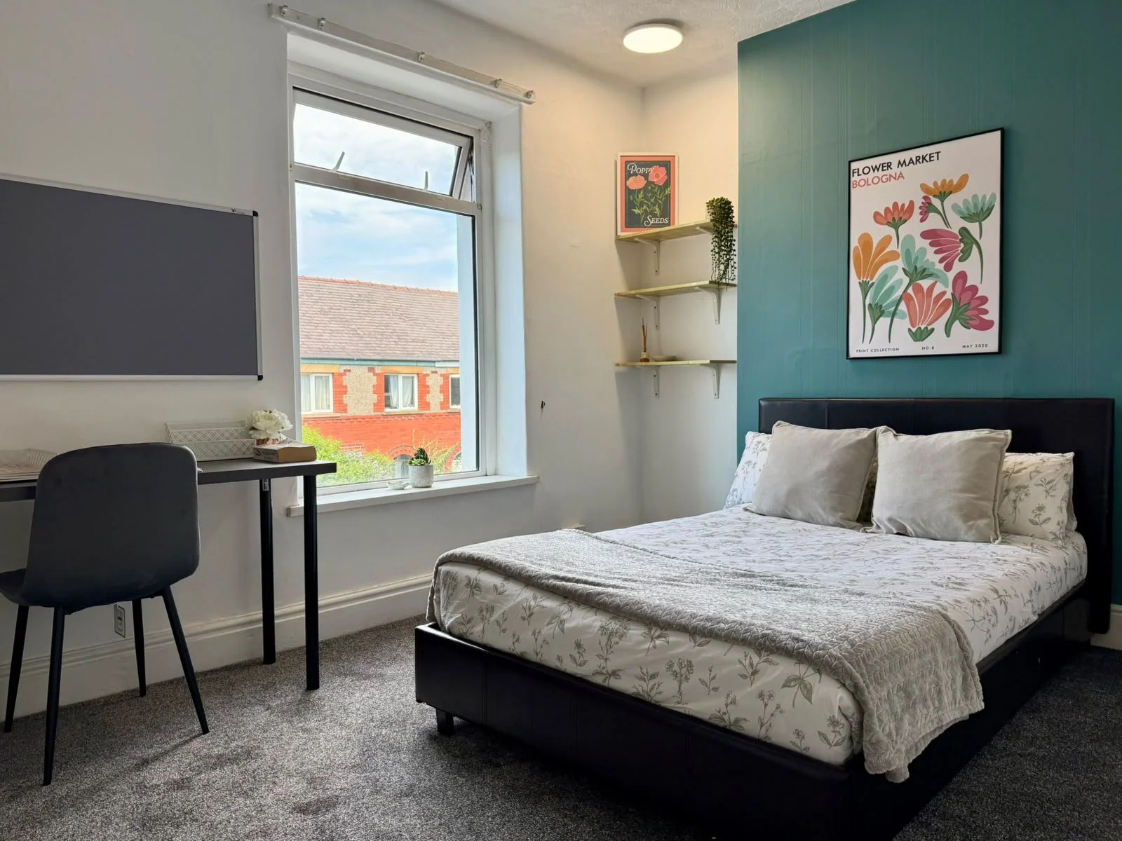 A bedroom with a bed, white floral bedding, and three pillows. There is a large window with a view of a red brick building outside. The wall behind the bed is painted teal and has a framed floral poster that says "Flower Market Bologna". To the left 