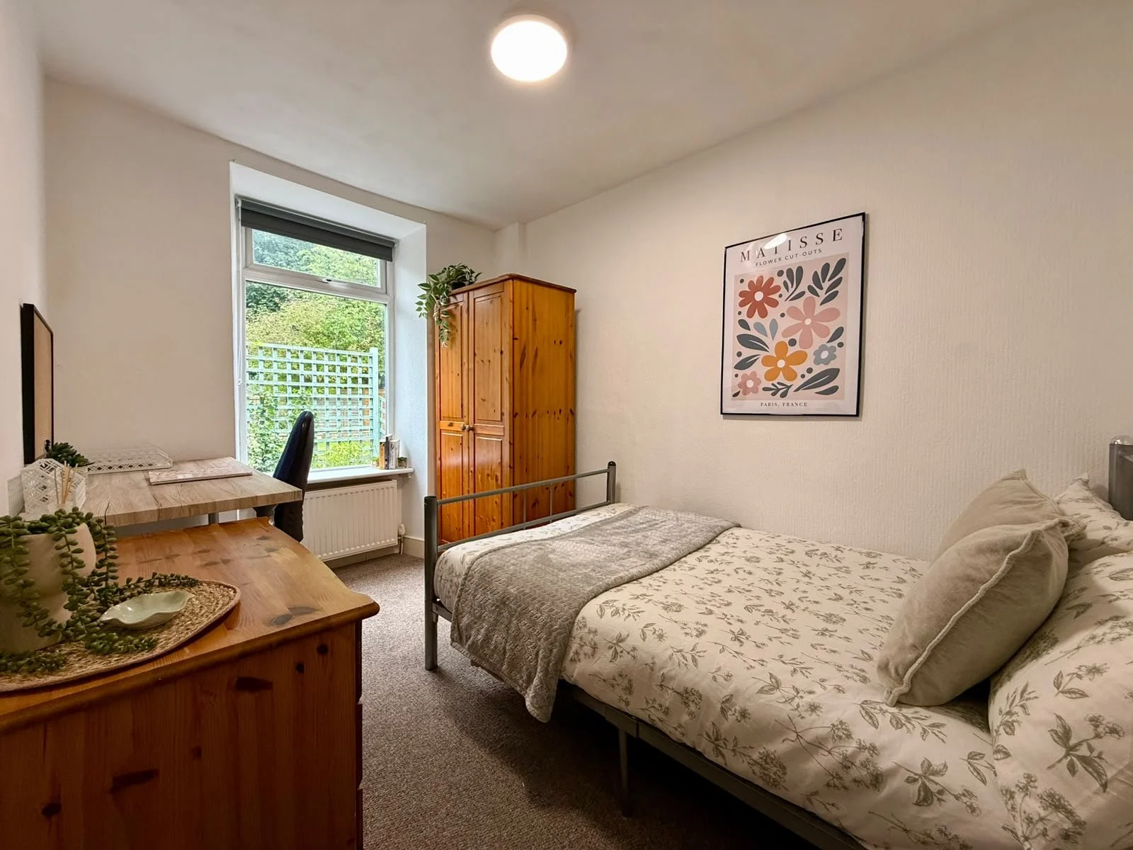 A cozy bedroom with a double bed, beige floral bedding, and pillows. A wooden wardrobe and a window with green view. A small desk with a black chair and decor items. Wall art featuring floral artwork. Carpeted floor and neutral walls.