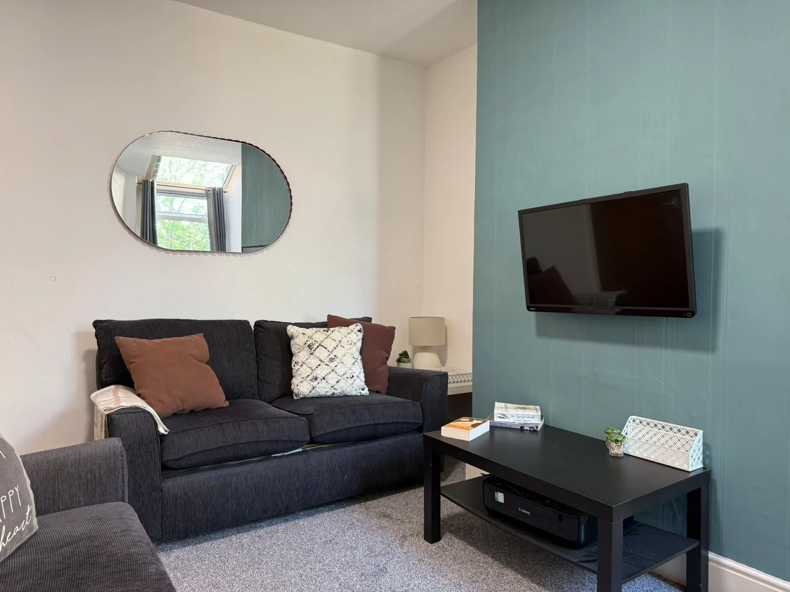 Living room with a black sofa, pillows, wall mirror, TV, side table, lamp, and decorative items.