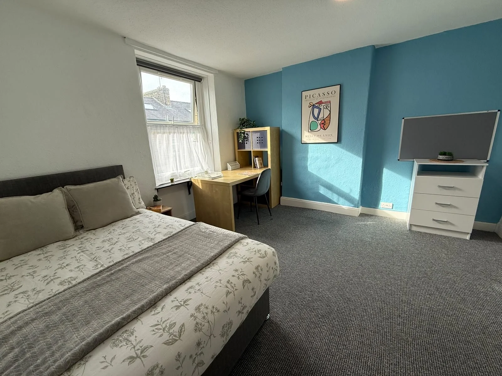 A bedroom with a bed, desk, chair, dresser with a TV on top, and a framed Picasso poster on a blue accent wall, with a window.