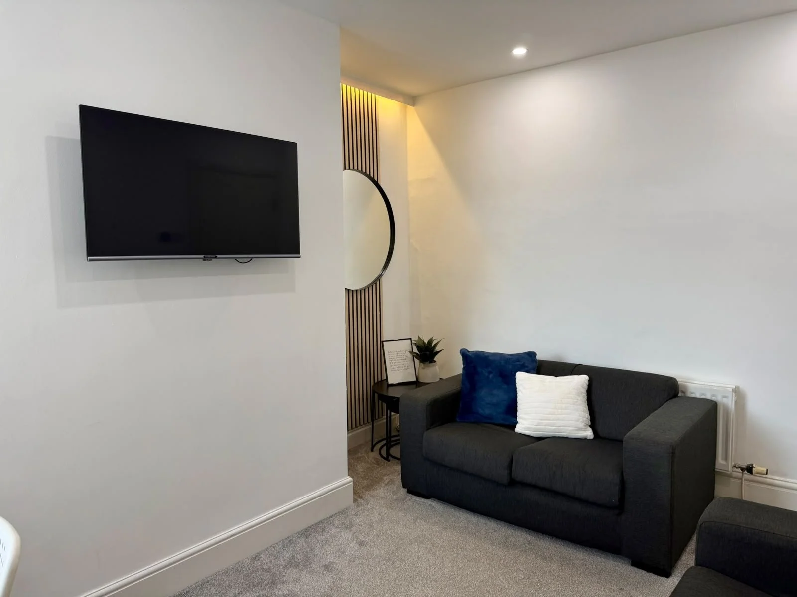 A living room corner featuring a wall-mounted flat-screen TV, a dark gray sofa with blue and white pillows, a small side table with a potted plant and framed picture, white walls, beige carpet, and a radiotor