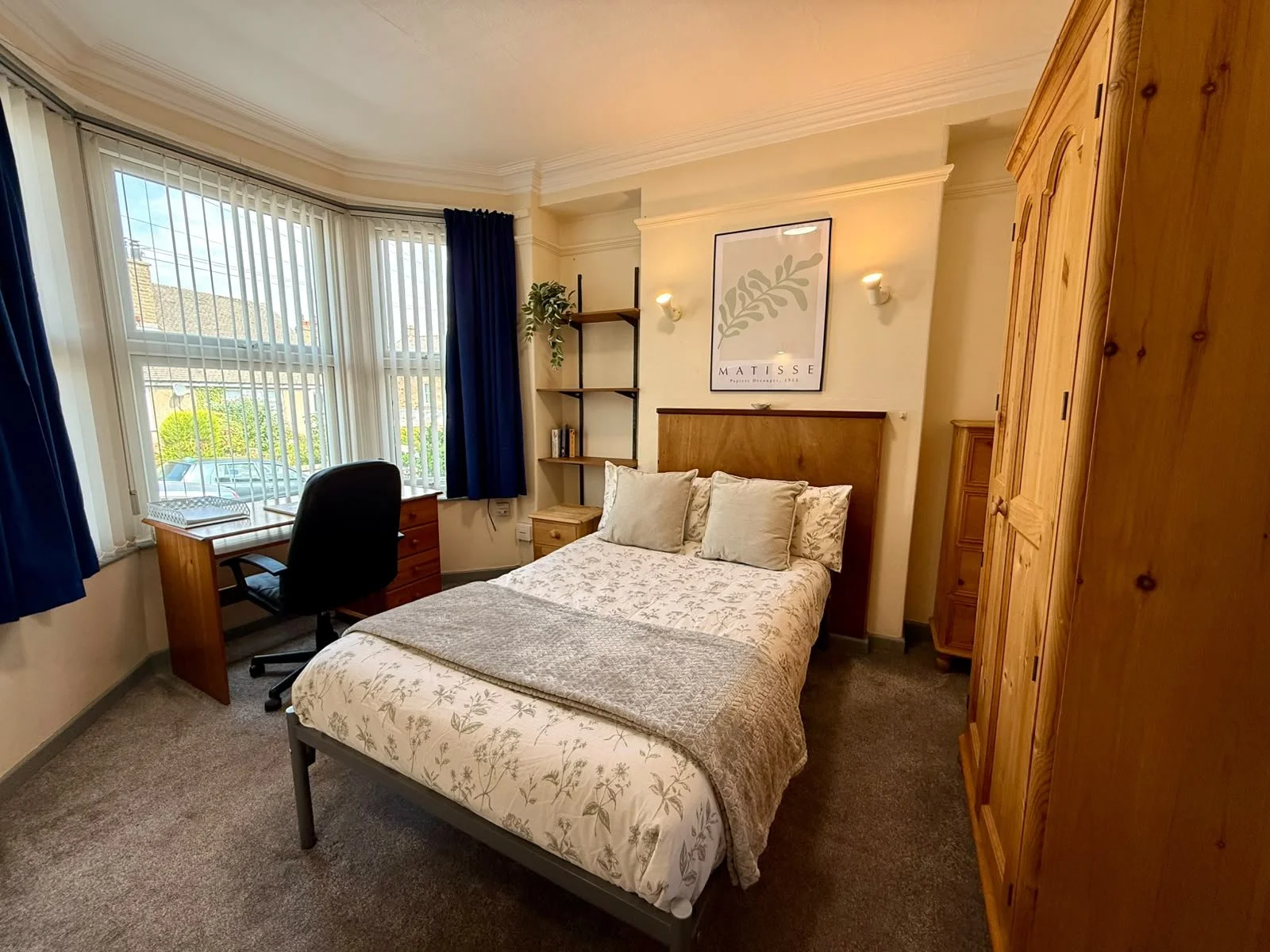 A cozy bedroom with a bed covered in a floral duvet and multiple pillows, a corner bookshelf with decorative plant, walls with artwork, a window with vertical blinds and blue curtains, a desk with a black office chair, a large wooden wardrobe, and ca