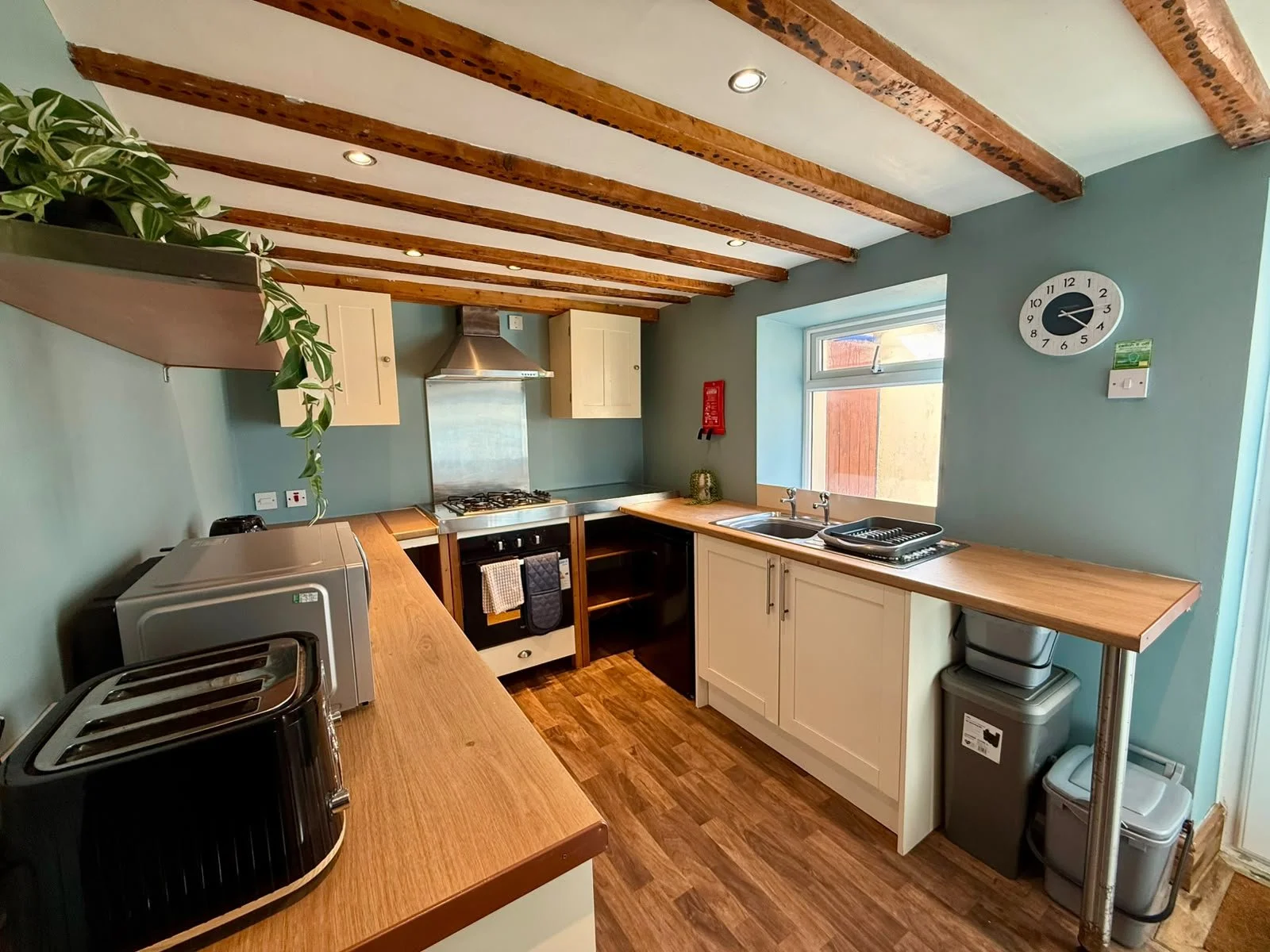 A small kitchen with wooden beams on the ceiling, teal walls, and white cabinets. Countertops hold a toaster, microwave, and dish rack. There's a window above the sink and a clock on the wall.