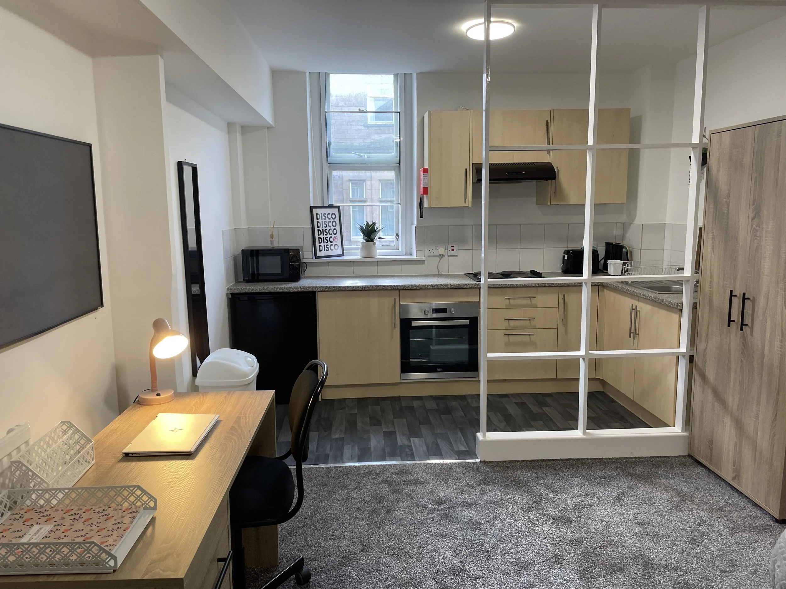 Interior of a small apartment unit with a bed on the right, a desk with a chair and a laptop, a kitchenette with wooden cabinets, a window, and some decorative art on the wall.