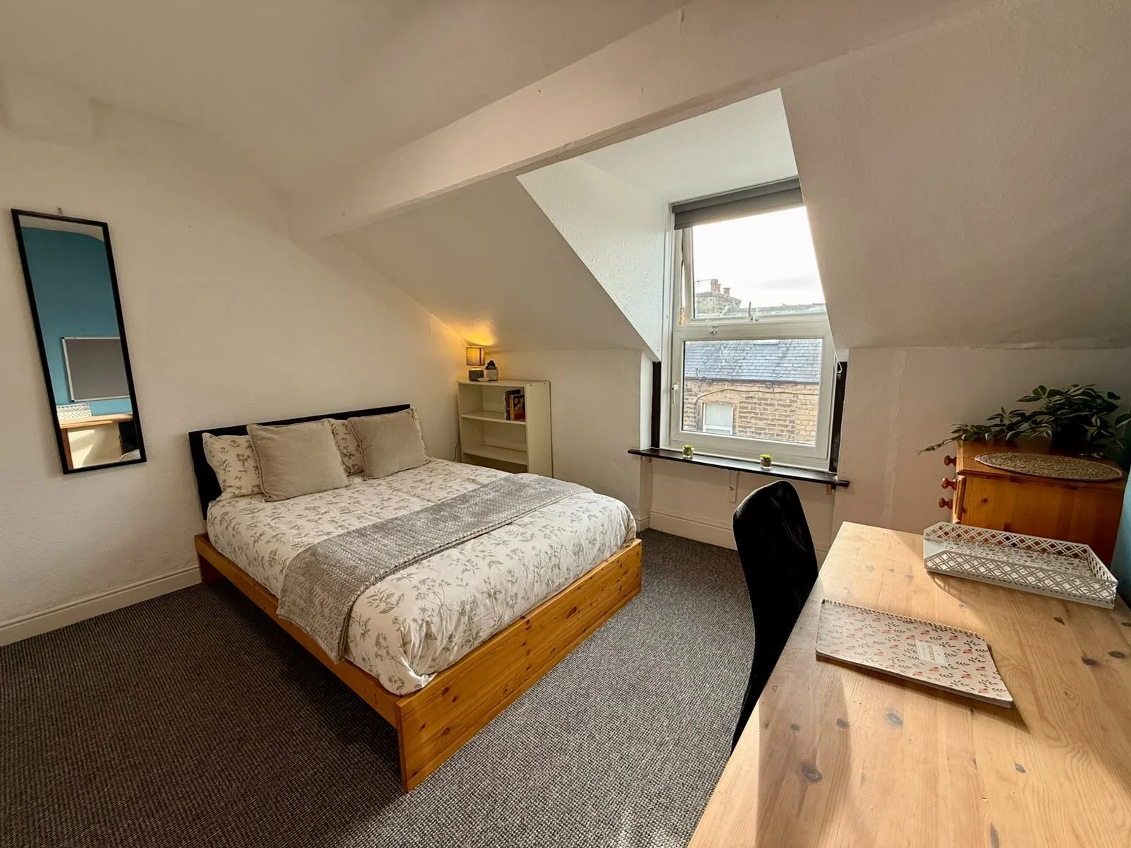 A cozy bedroom with a double bed, white bedding, and beige pillows, next to a large window with a view of neighboring buildings, a small wall mirror, a white bookshelf with decor, and a wooden desk with a plant and a magazine.