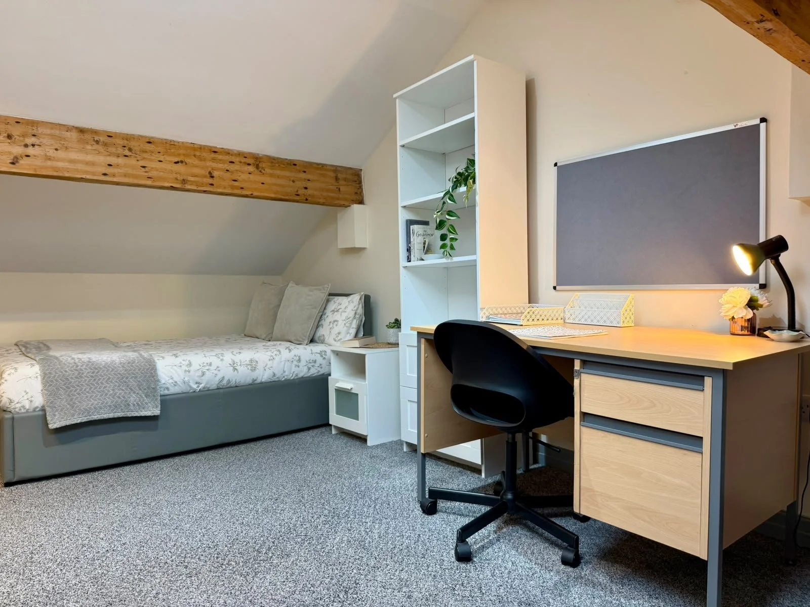 A bedroom with a single bed, a small white nightstand, a white bookshelf, a desk with a black chair, a green plant, and a gray bulletin board on the wall. The room has a sloped ceiling with a exposed wooden beam.