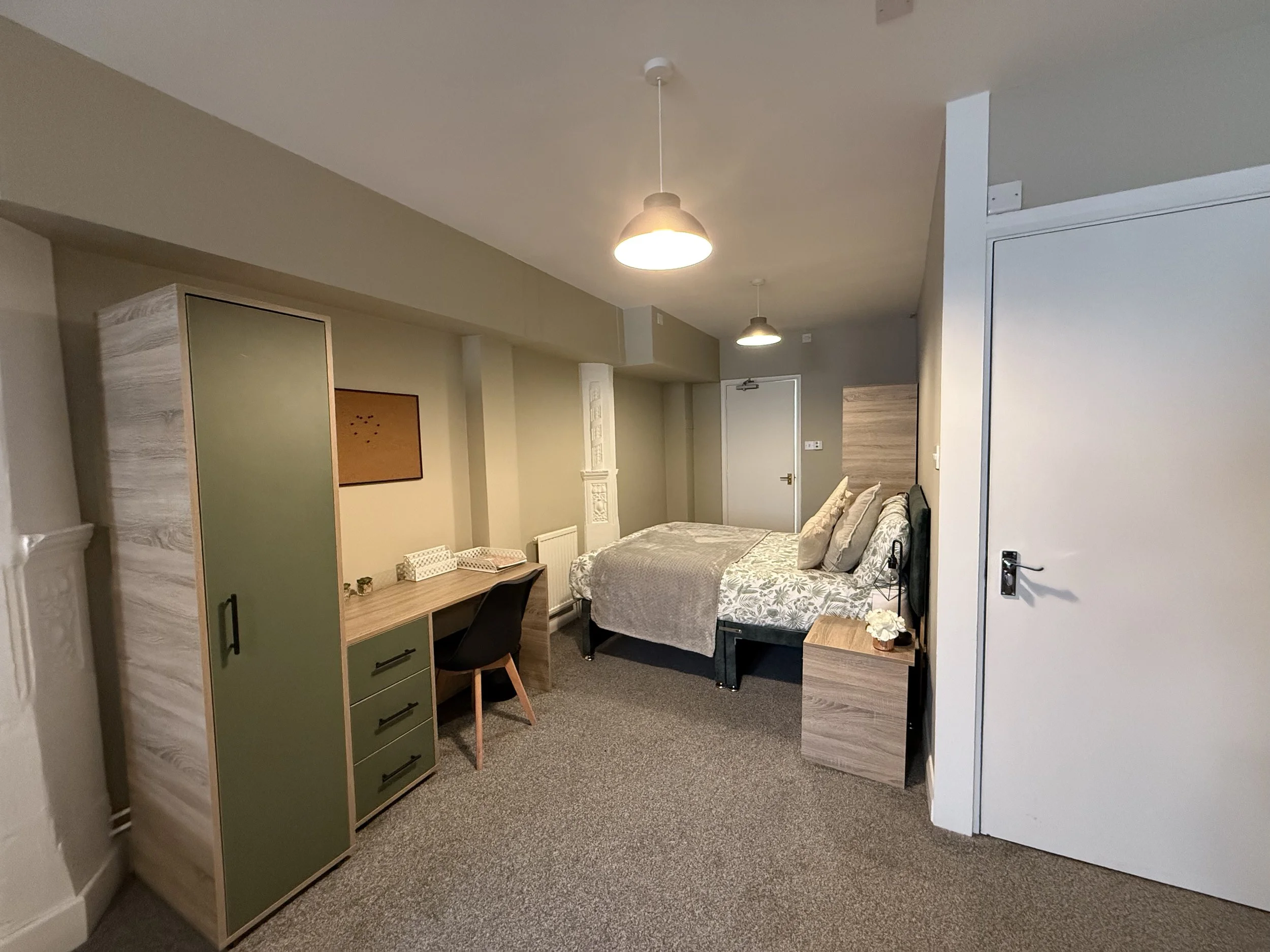 A modern bedroom with a bed in the center, a nightstand with a flower, a desk with a chair, a wardrobe, and hanging ceiling lights.
