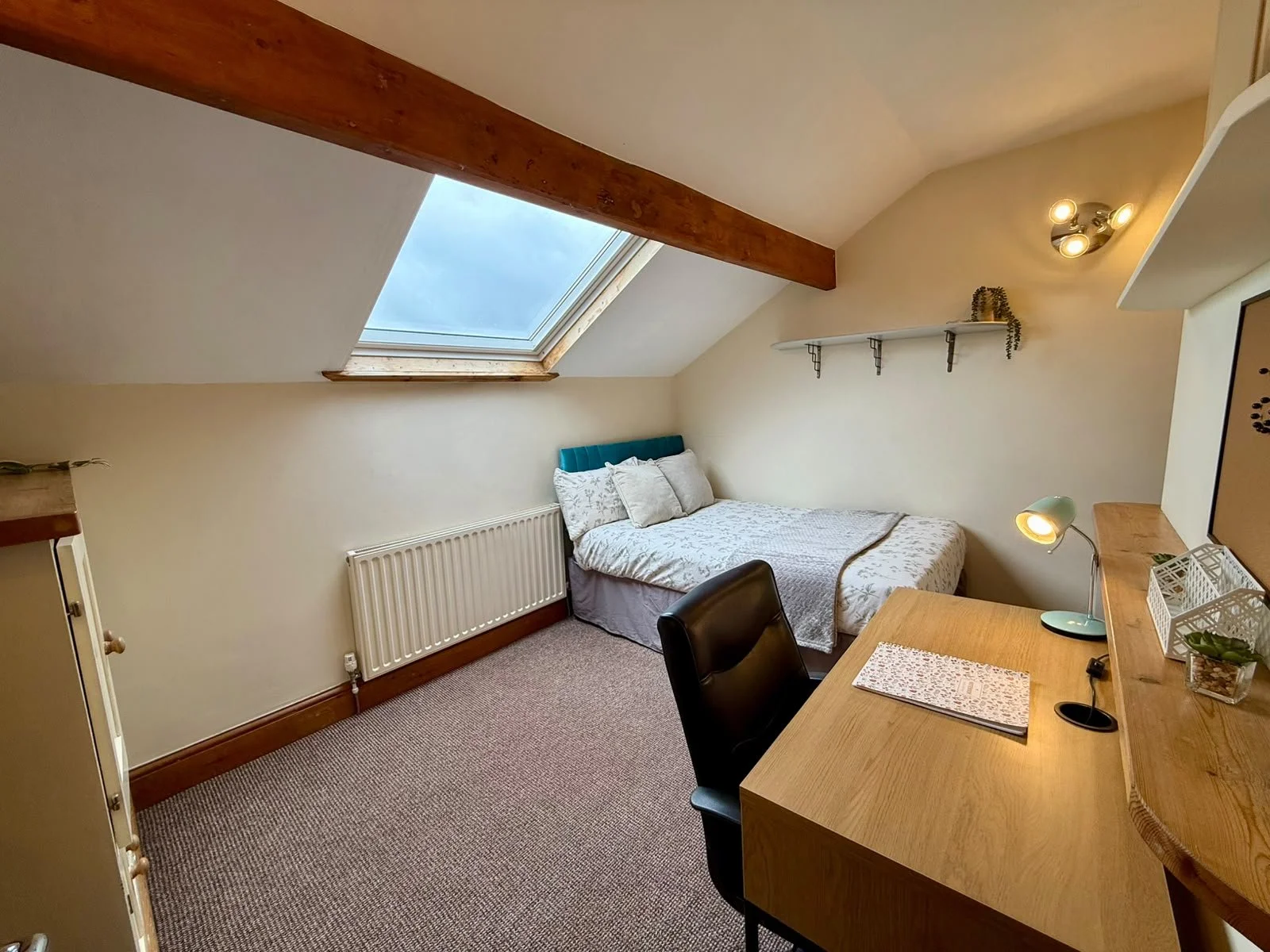A cozy bedroom with a sloped ceiling and a skylight. The bed has white linens and pillows, positioned against the wall beneath the skylight. There's a wooden desk with a black chair, a small desk lamp, and some decorative items. A shelf with plants i