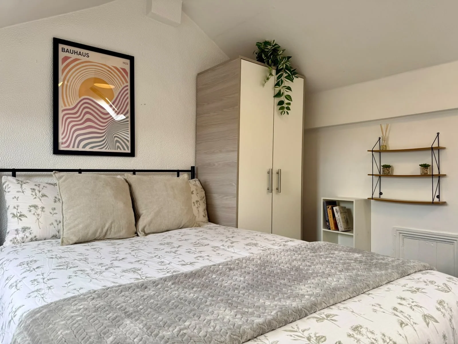 A cozy bedroom corner with a bed featuring a botanical patterned bedsheet and beige pillows, a black framed Bauhaus art poster on the wall, a white wardrobe with wood accents, a small white bookshelf with books, and a wall-mounted wooden shelf with p