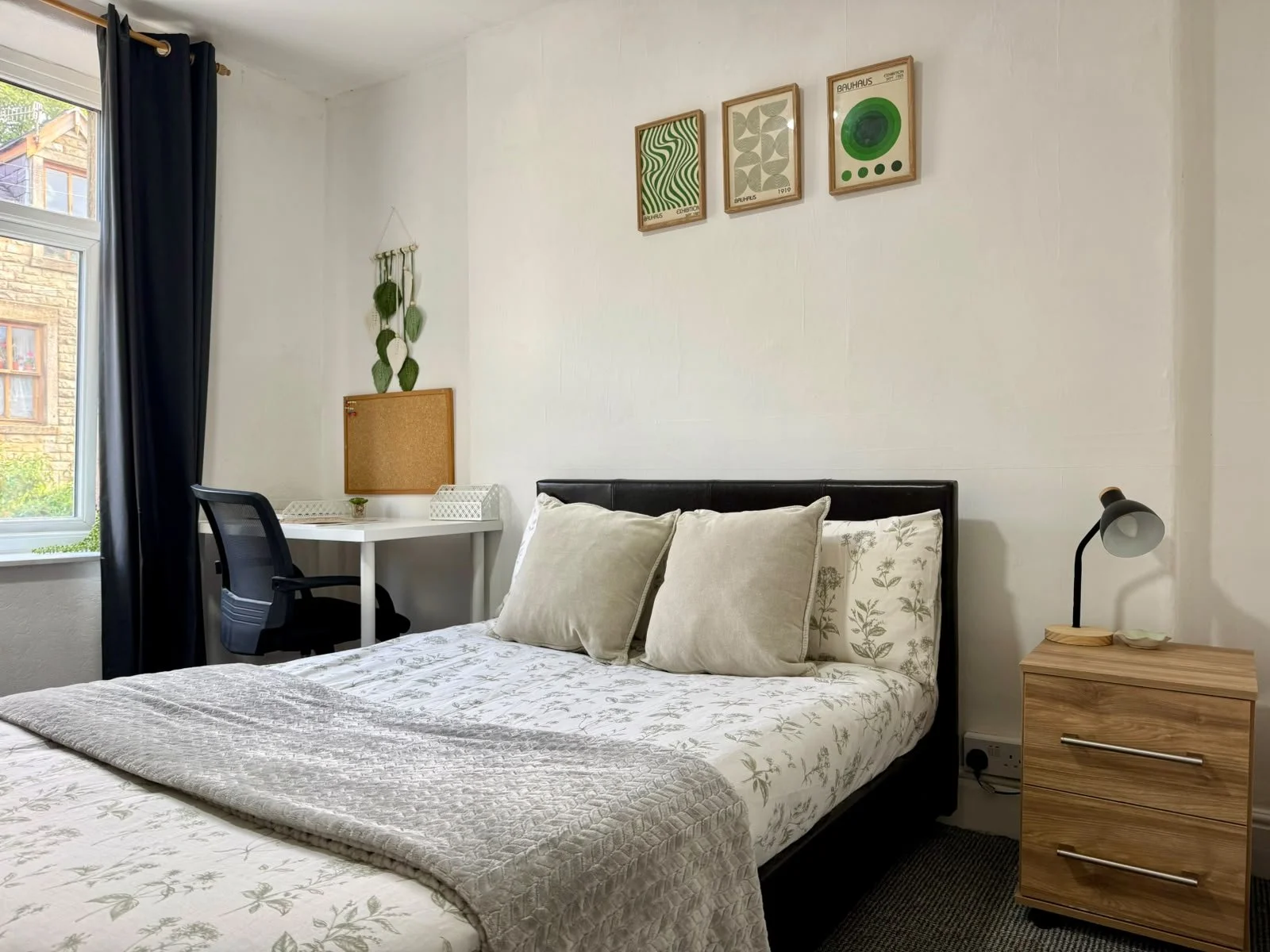 A cozy bedroom with a bed featuring a floral quilt and pillows, a nightstand with a lamp, a desk with a chair by a window, and three framed art prints on a white wall.