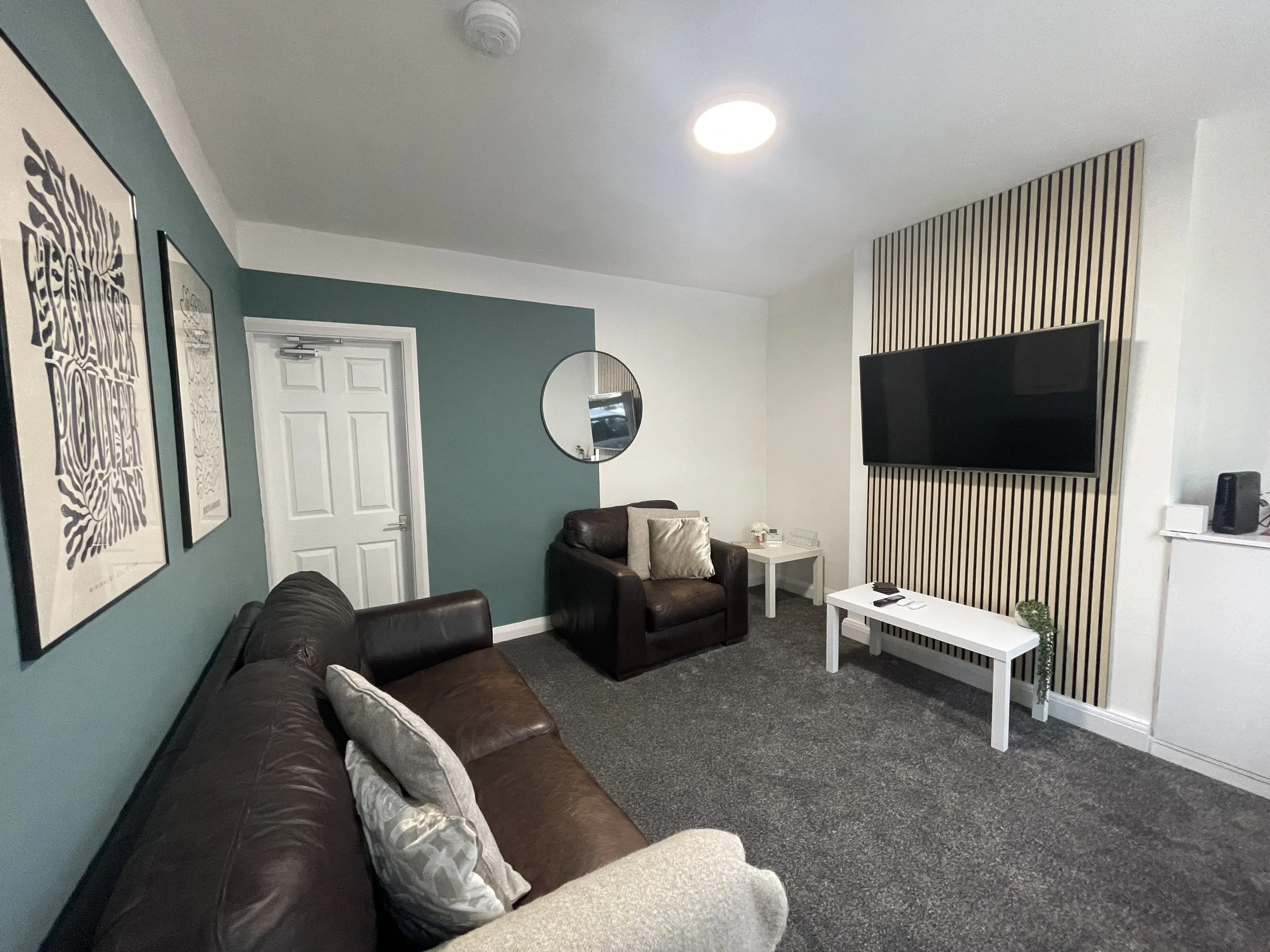 Living room with a brown leather sofa and matching armchair, teal accent wall with framed artwork and a round mirror, white side table, large flat-screen TV mounted on a striped wood panel, and a white coffee table on a gray carpet.