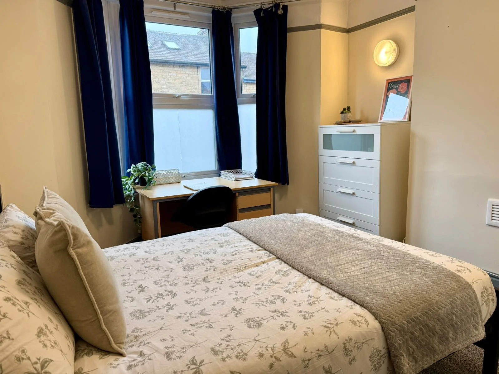 A cozy bedroom with a bed covered in floral bedding, beige pillows, and a beige quilt. There is a small desk with a black chair and green plants near the window. The window has dark blue curtains and shows neighboring rooftops. A white dresser with a